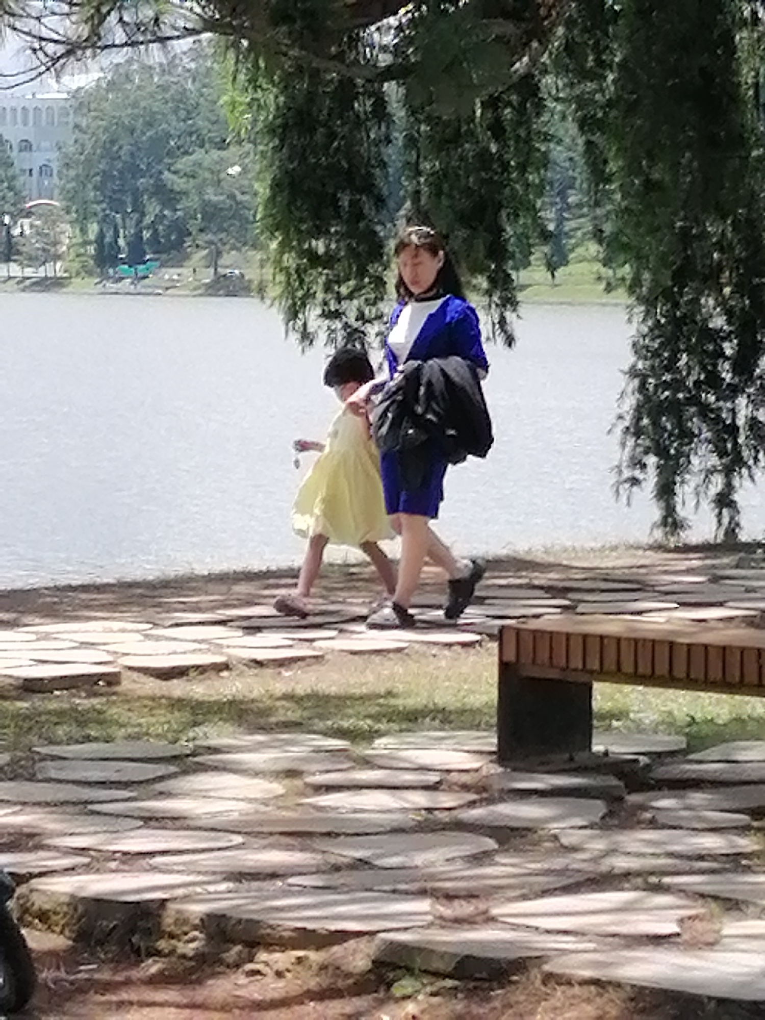 A young girl in a yellow dress and a woman in a blue jacket and white shirt walk along a stone pathway beside a lake. The girl is on the left, and the woman is on the right. The woman is carrying a black bag over her shoulder. They are walking away from the camera. The pathway is made of irregularly shaped flat stones laid on the ground. In the background, there is a body of water, trees, and a building. The scene appears to be in Da Lat, Vietnam, likely near Xuan Huong Lake, given the presence of a lake, trees, and the general ambiance.