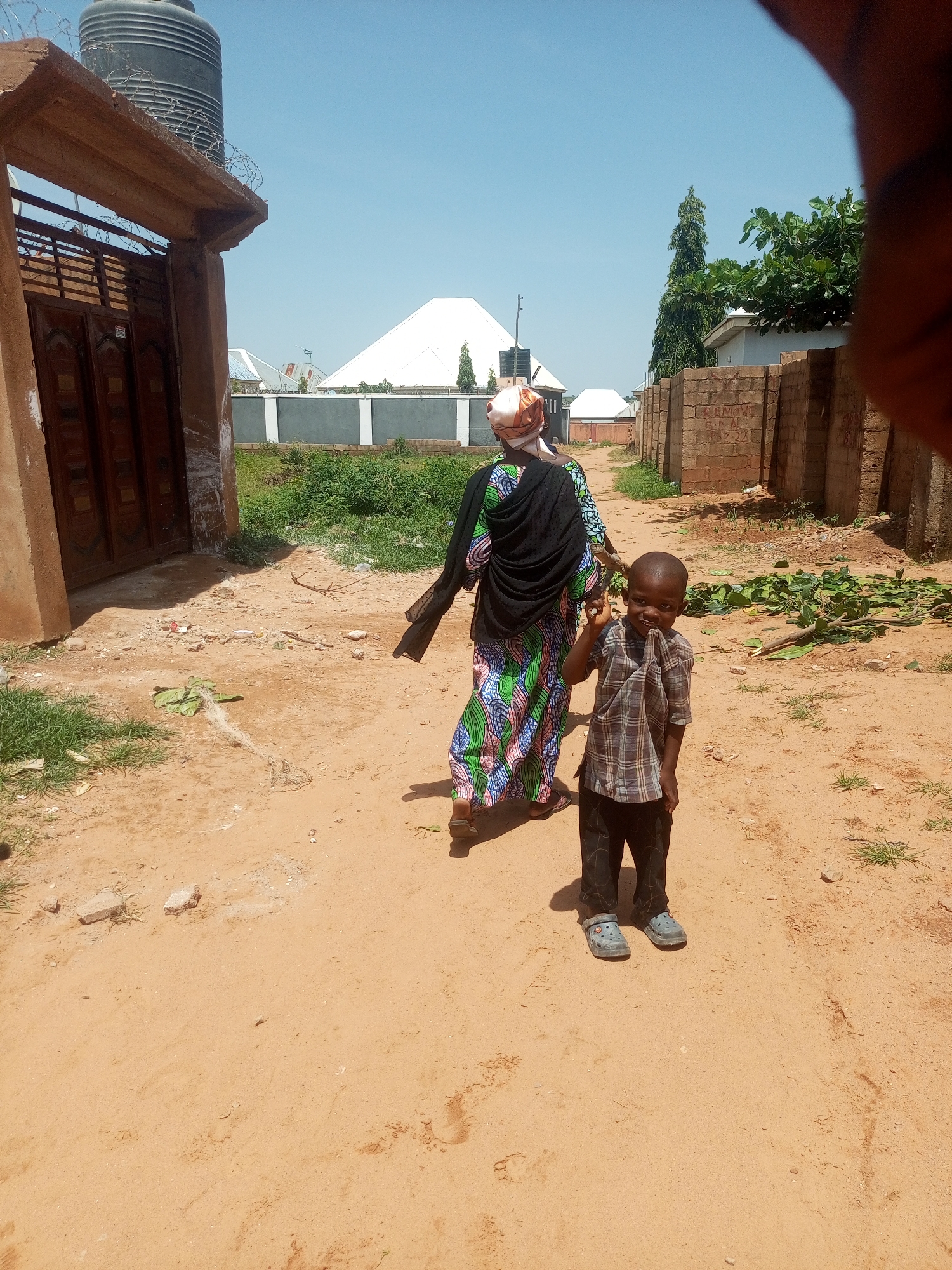 In Tempure, Nigeria, a young Black boy in a checkered shirt and dark pants stands on a dirt road, smiling and raising his right hand. He wears grey clog-style shoes. Behind him, a Black woman walks away from the camera, wearing a patterned dress and a dark headscarf. She is carrying a dark cloth over her left arm. The environment is dusty and dry, with scattered debris and some sparse vegetation. A building with a brown wooden door is to the left, and a brick wall and more buildings are visible in the background. A large water tank sits atop a structure to the left. The sky is clear and blue.