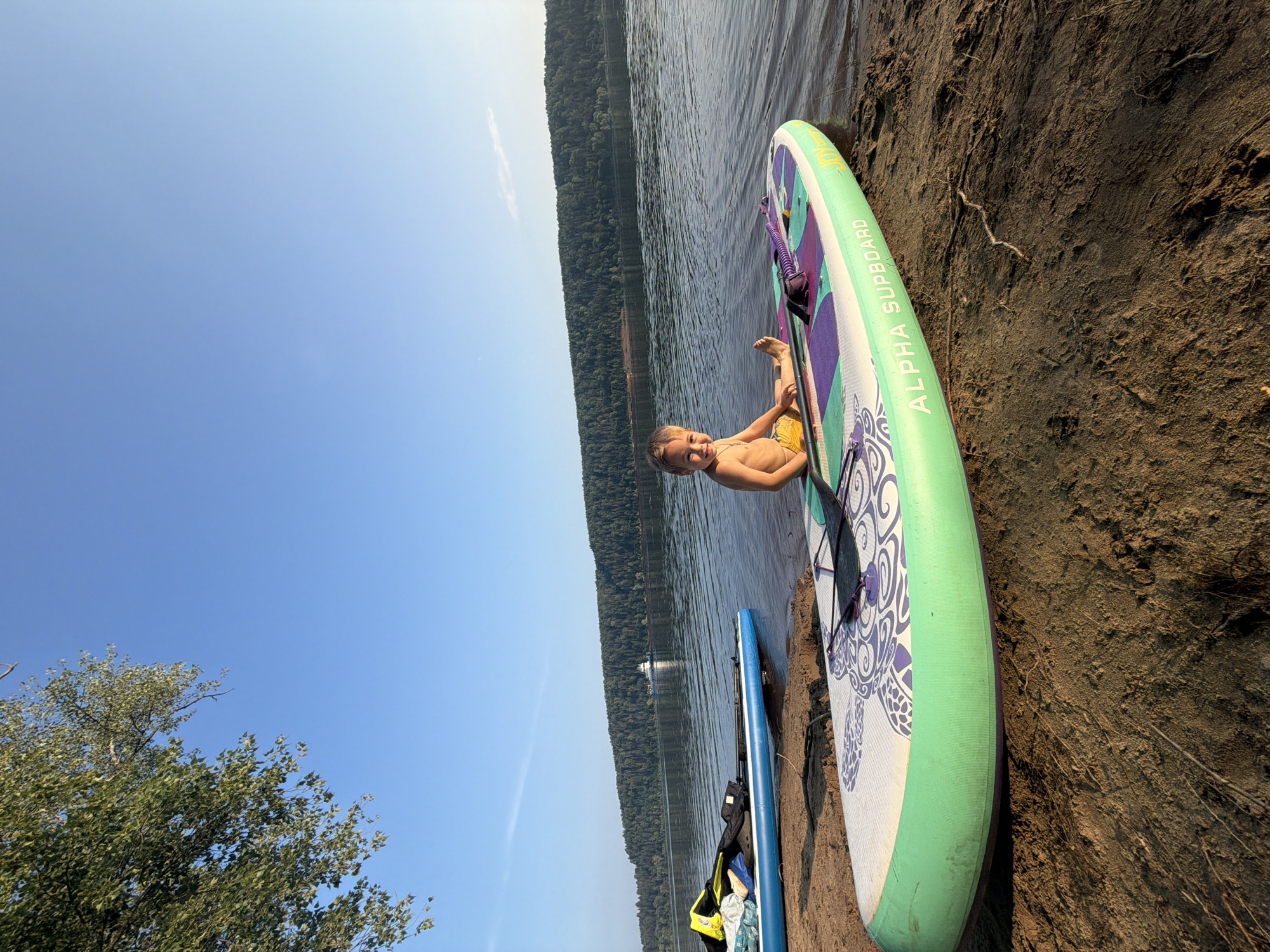 A young boy with blond hair and yellow shorts sits on a green and purple paddleboard on a sandy shore. The paddleboard has the words "ALPHA SUPBOARD" written on its side. Behind the boy, a body of water reflects the clear blue sky. In the distance, a dense forest lines the opposite bank of the water. Another blue paddleboard with some gear on it is visible to the left of the boy. A boat or ferry is seen on the water in the background. The scene appears to be a calm summer day by a lake or river, possibly in the vicinity of Ol'khovskoye, Russia, given the description of the location. The lighting suggests it is either early morning or late afternoon.