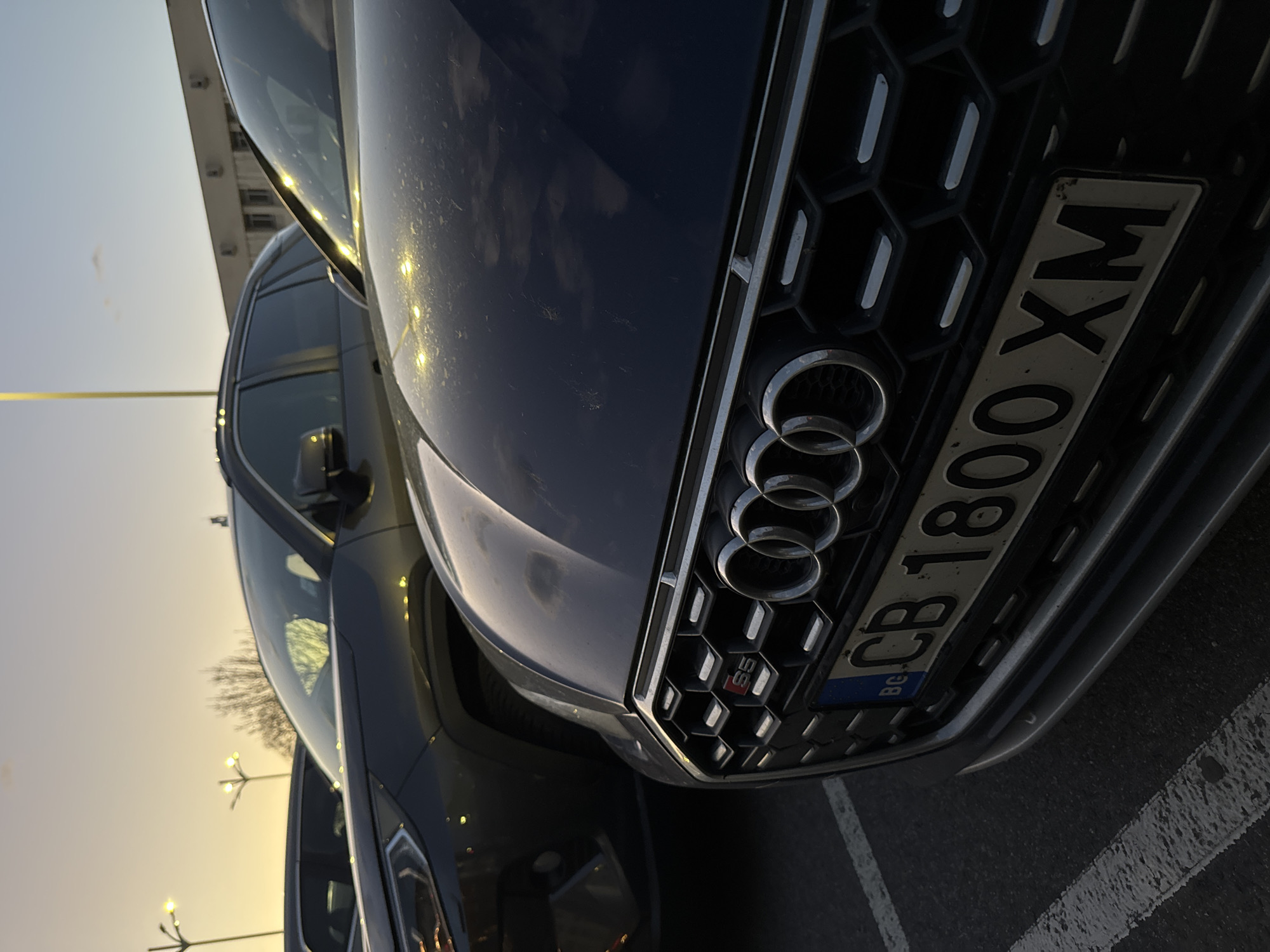The image shows a dark-colored Audi S5 with a Bulgarian license plate "CB 1800 XM". The vehicle is parked in a parking lot, possibly in Plovdiv, Bulgaria, although there is no explicit confirmation of the location within the frame. The sky suggests either sunrise or sunset. Other vehicles are partially visible in the background. The focus is on the front of the Audi, highlighting the grille, the Audi rings logo, and the S5 emblem. The lighting is slightly uneven, with some bright spots on the car's hood.
