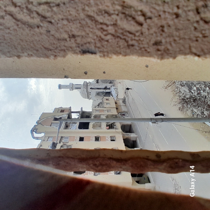 The image displays a war-torn building and mosque through a gap in a ruined structure. The building's facade is severely damaged with gaping holes. A mosque with a slender minaret stands in the background, also showing signs of destruction. Debris litters the street, and two dark figures appear to be moving along the road. The shot is taken from a low angle, creating a sense of claustrophobia and highlighting the desolation. The sky is overcast, intensifying the somber atmosphere.