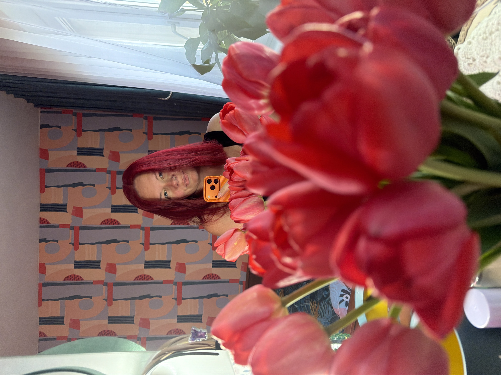 A woman with red hair, wearing a black top, takes a selfie with an orange phone against the reflection in a mirror. In the mirror, one can see a patterned wall, and part of a white curtain and window. A vase filled with red tulips is in the foreground, obscuring some of the reflection. The location is implied to be in Chaikovsky, Russia.