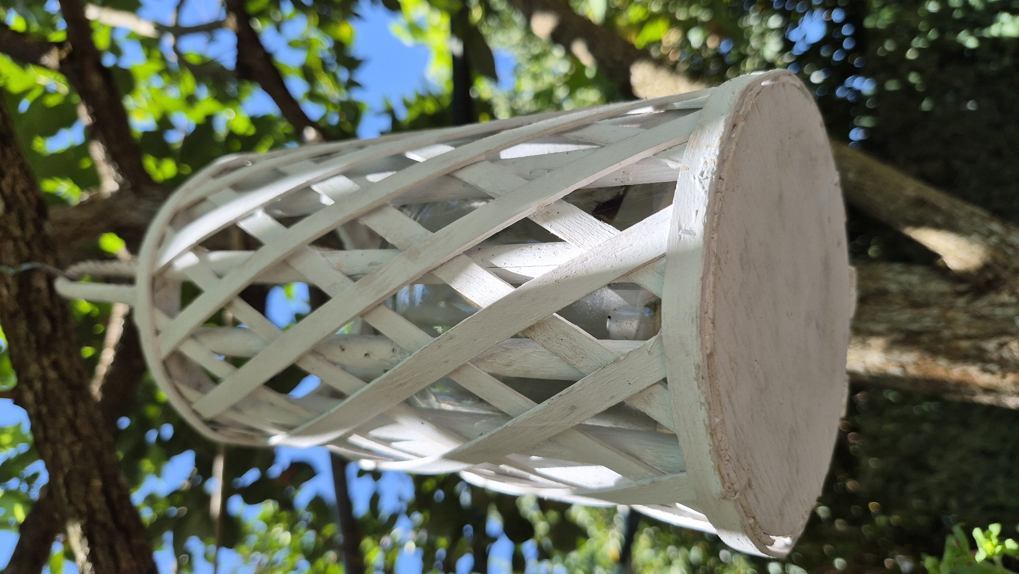 A white, lattice-style lantern is hanging from a tree branch, against a backdrop of green leaves and a blue sky. The lantern is cylindrical with a round base and top. The lattice pattern is made of thin wooden strips that crisscross each other. Inside the lantern, a glass jar is visible. The tree branches and leaves are out of focus, suggesting a shallow depth of field. The lighting indicates it is daytime, with sunlight filtering through the leaves.