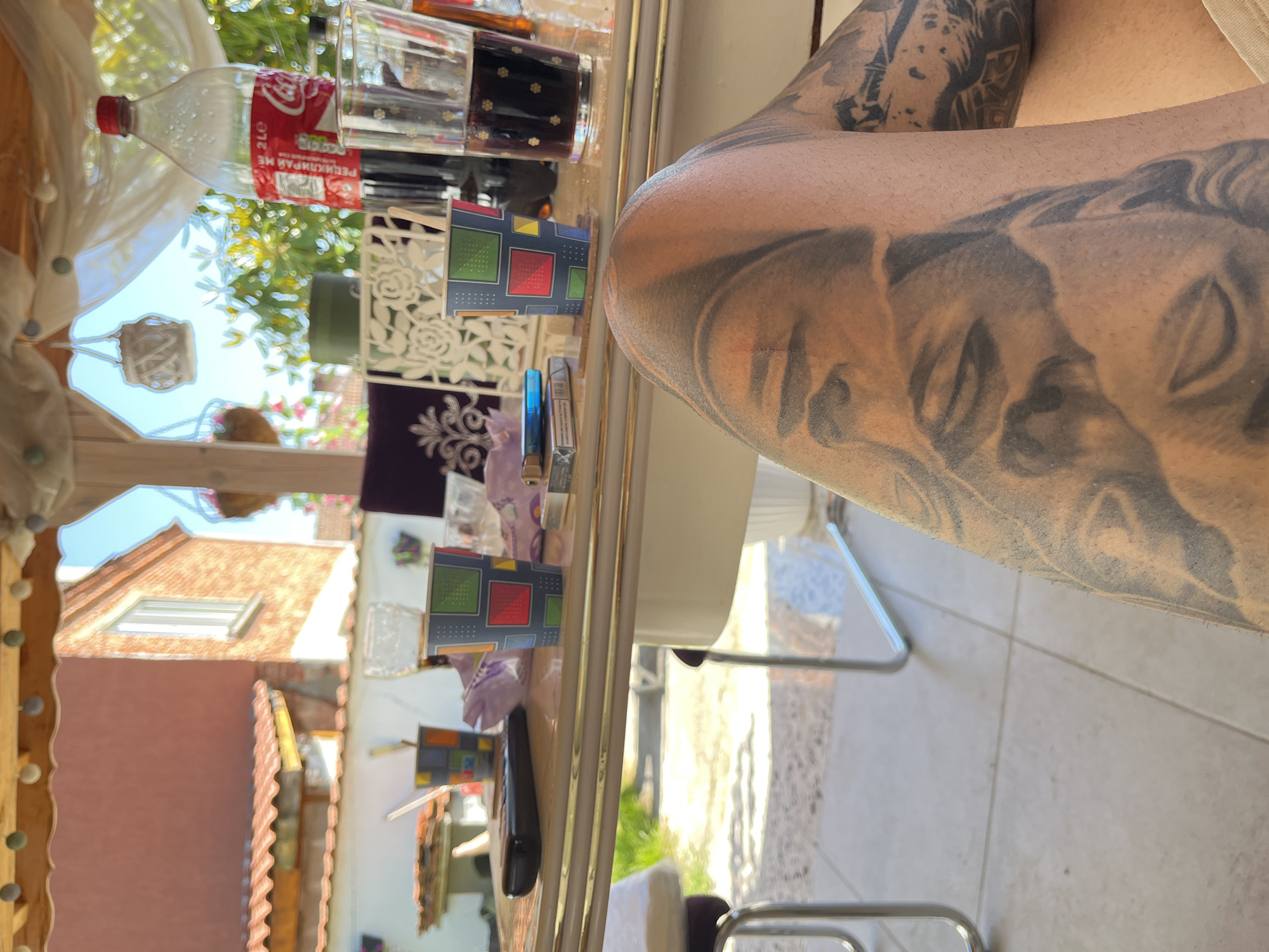 The scene depicts a table setting with various objects, likely outdoors, possibly in an area outside a residence. A bottle of Coca-Cola, a glass filled with a dark liquid, and two small decorative boxes are positioned on the table. Also present are cigarettes, lighters, and several glasses. Part of a tattooed human arm is prominently featured. The background shows a building with a terracotta tile roof. The location appears to be a residential area, based on the presence of the building and the style of the outdoor setting.