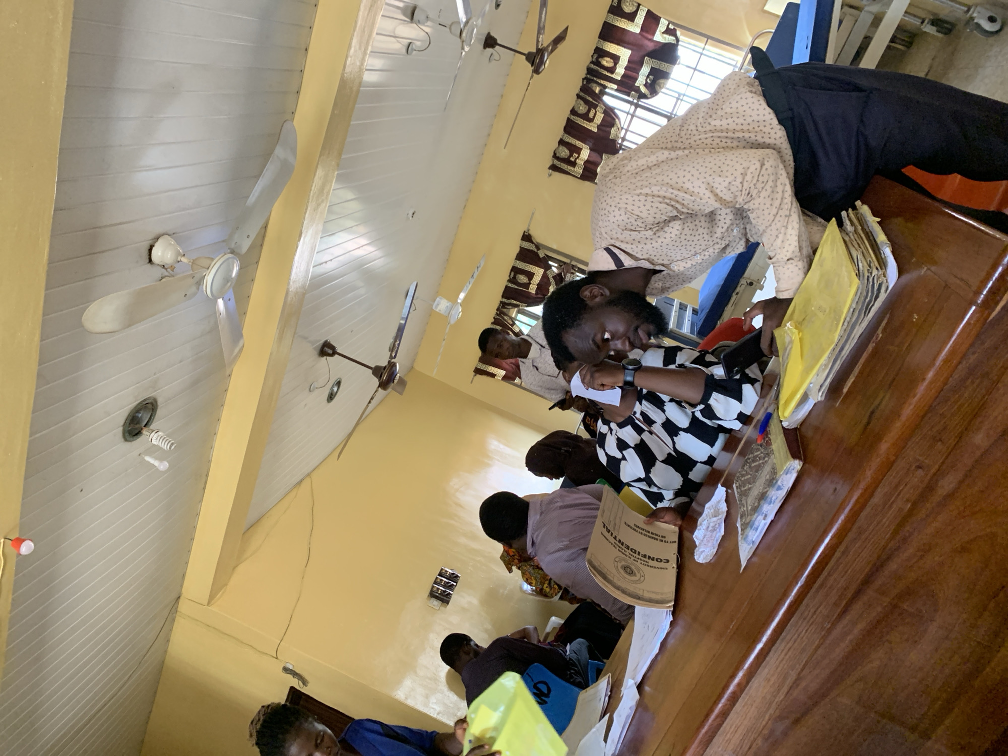 The image depicts an indoor scene in Oko Erin, Nigeria, likely a classroom or office setting. Several individuals are seated around a long wooden table, engaged in what appears to be a meeting or review of documents. One man, wearing a patterned shirt and dark pants, is leaning over the table, possibly pointing at or examining papers. Others at the table are looking down at documents, some holding pens or writing. The documents on the table include folders labeled "CONFIDENTIAL". The room features a white ceiling with ceiling fans and yellow-painted walls with windows covered by patterned curtains. A dangling light fixture is visible.