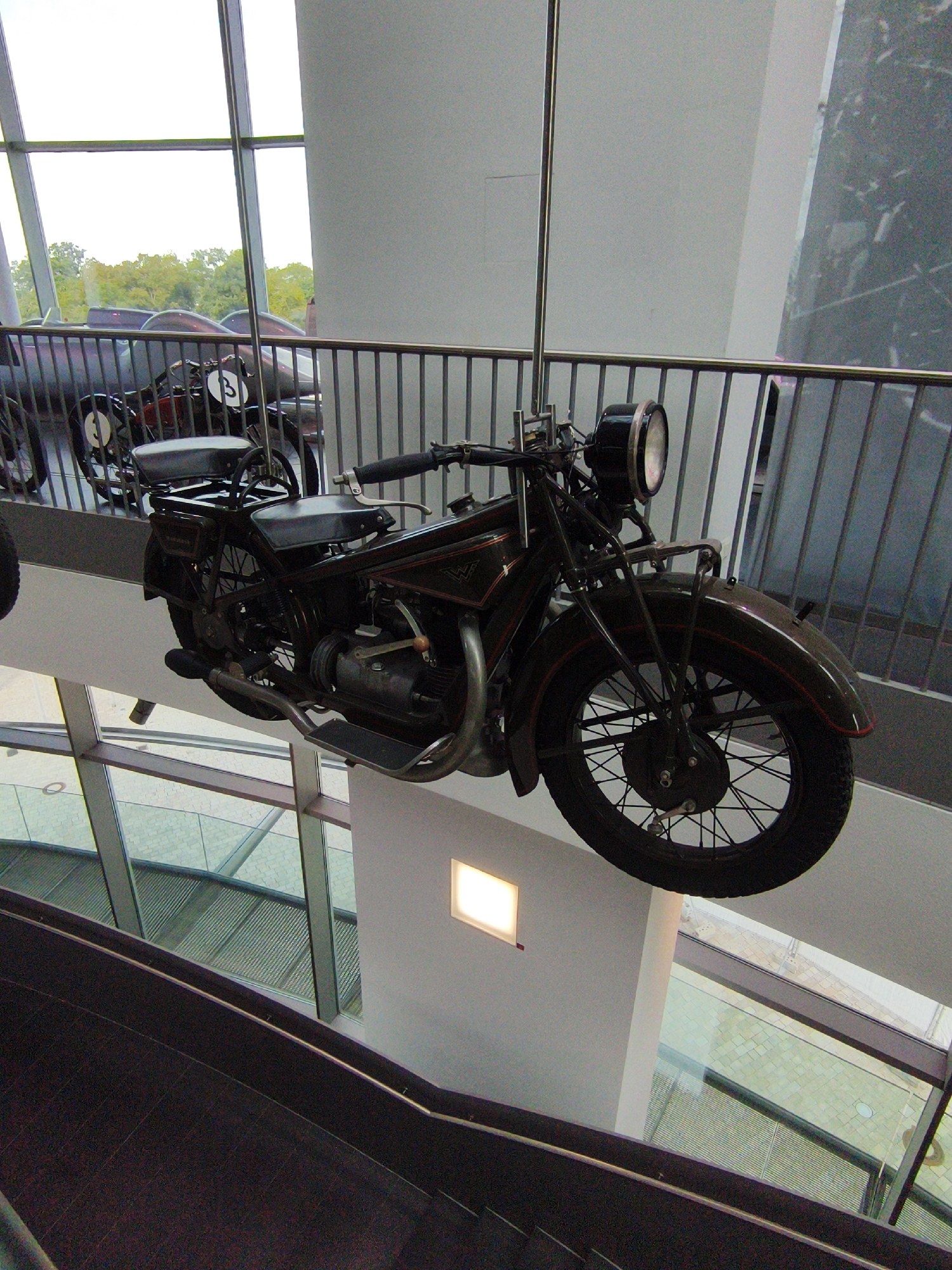 The image shows a vintage motorcycle suspended in the air, likely as an exhibit. The motorcycle is dark colored with chrome accents and has a classic design. It is positioned in front of a metal railing and a large window, suggesting an indoor exhibition space. The background reveals an outdoor scene with trees and a bright sky. The interior space is modern, with white walls and visible structural elements. The overall impression is one of a well-lit museum or gallery. This photograph was taken in Ingolstadt, Germany.