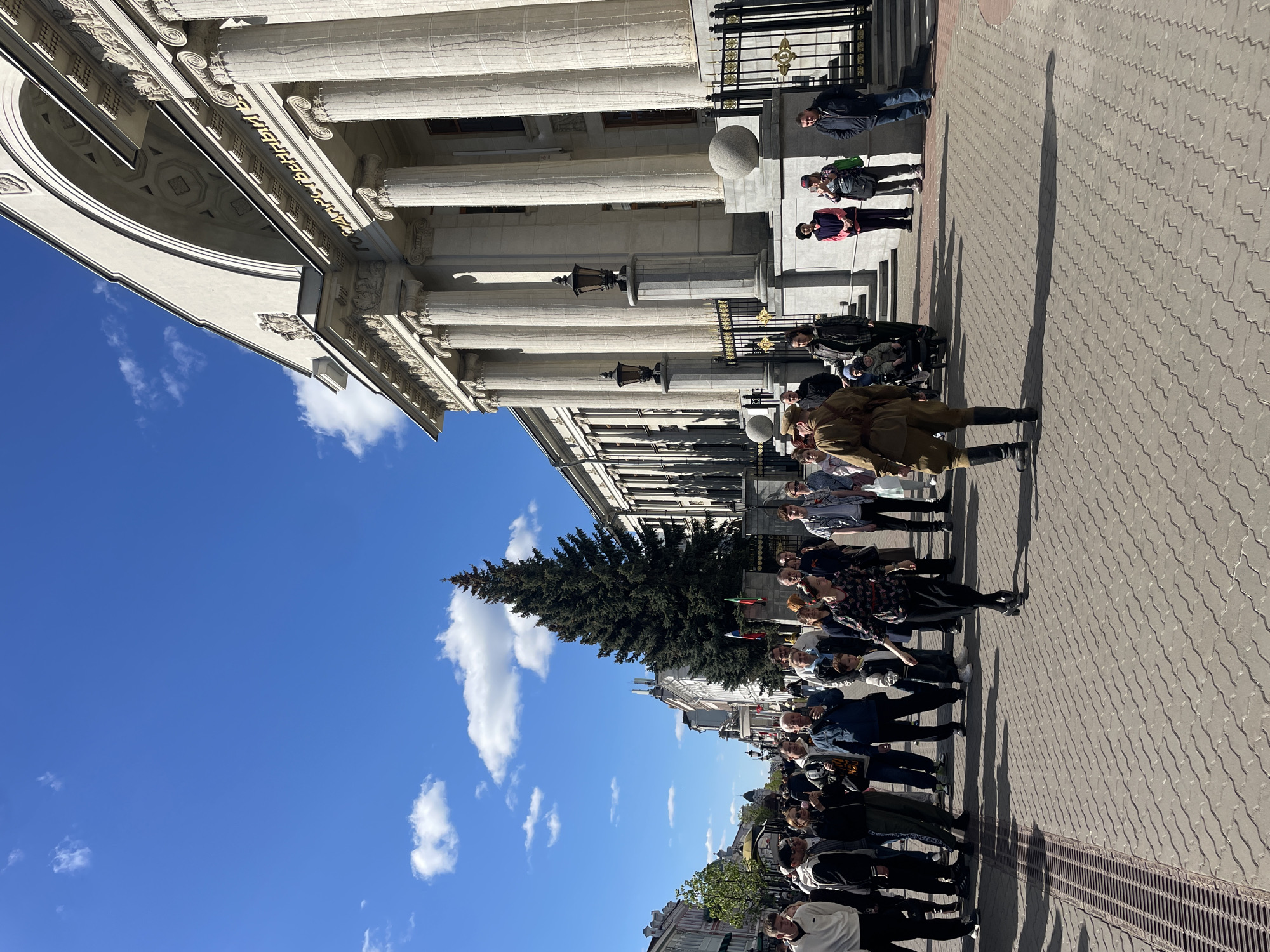 The image shows a group of people gathered on a paved walkway in front of a classical-style building with columns. The building appears to be located in Kazan, Russia.
Several people are standing in a line, while others are in the background. One person stands out as they are dressed in a military-style uniform from the World War II era. A large evergreen tree stands nearby, partially obscuring some of the building.
The sky is blue with scattered white clouds. The lighting indicates a sunny day. The scene suggests a public gathering or event of some kind, possibly commemorating a historical event.