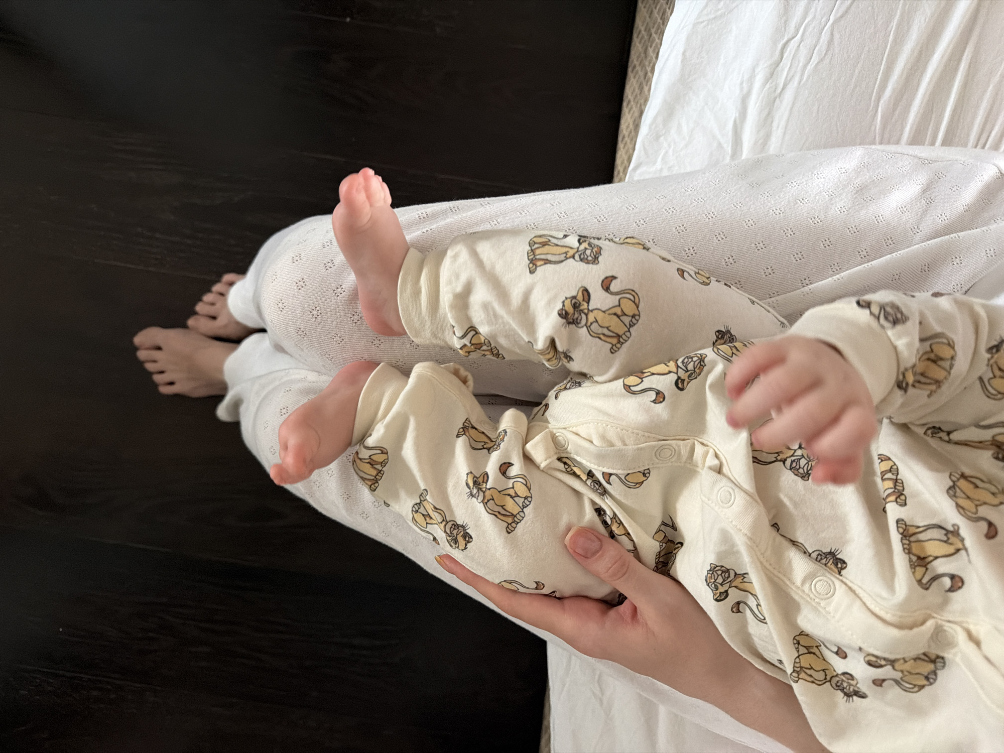 A photograph shows a person holding a baby wearing a light-colored, printed onesie. The person is wearing white pants and a textured top. The baby's bare feet are visible. The person's bare feet are also partially visible. The background includes dark wooden flooring and white bedding. There is no visual indication that the location is Khimki, Russia.