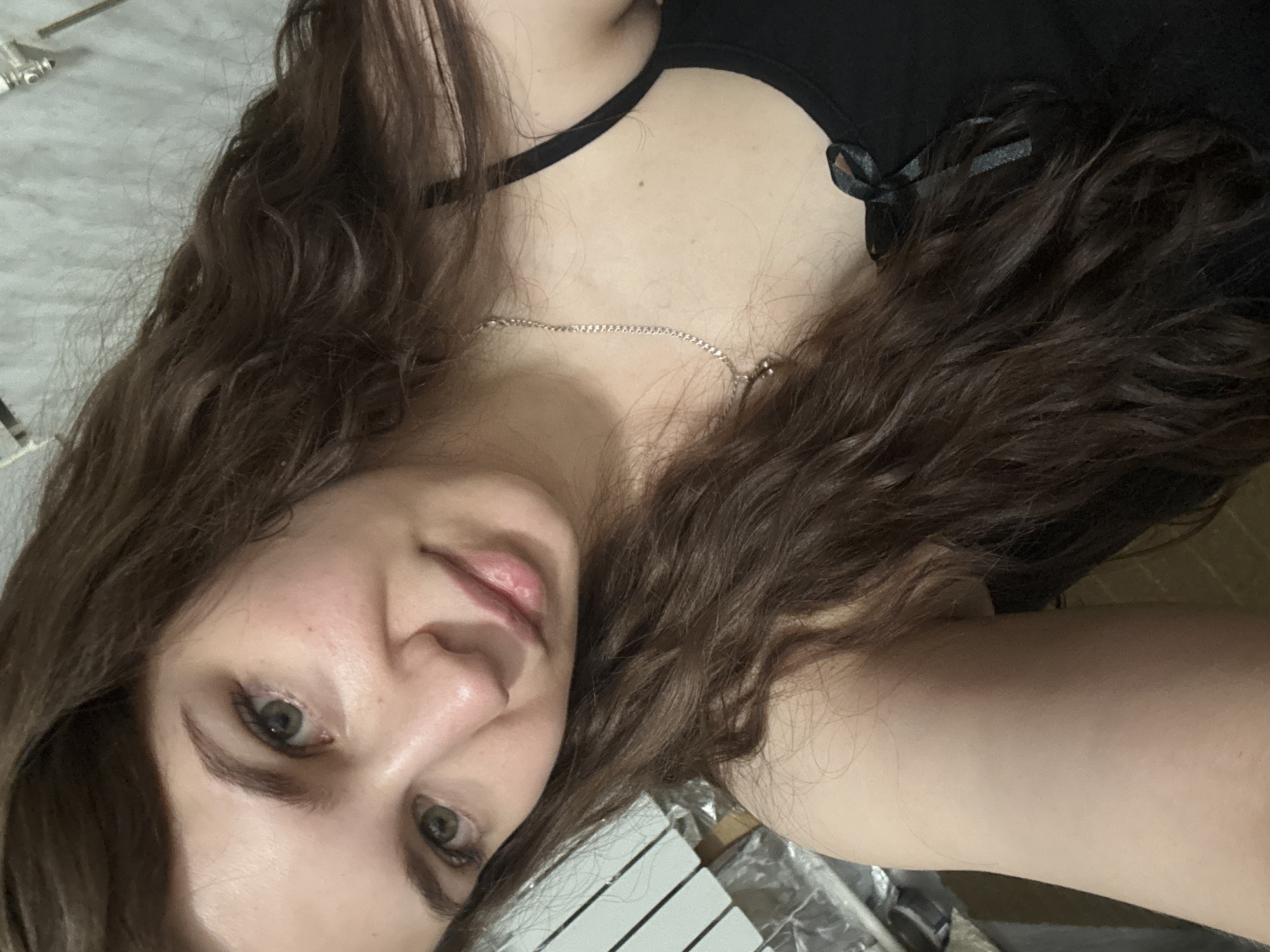 A woman with long, wavy brown hair is shown in a selfie. She has fair skin and green eyes. She is wearing a black top with spaghetti straps and a small bow. A silver necklace is visible around her neck. The background is indistinct, but some white and silver objects are visible in the lower right corner. The location of Nizhnyaya Gar', Russia is not evident in the image.
