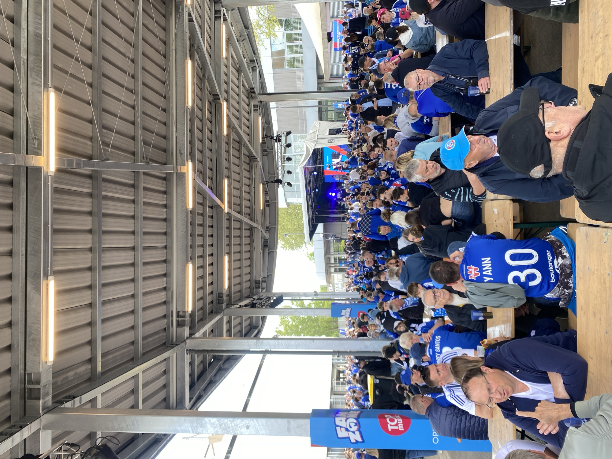 The image shows a large gathering of people, mostly dressed in blue, suggesting they are fans of a team or attending an event with a blue theme. They are seated at long rows of tables and benches, indicating an outdoor event or a covered area. In the background, there is a stage set up with a screen, hinting at a presentation or performance. The structure overhead appears to be a metal framework with lighting fixtures. Based on the clothing and the "TOP Music" branding, this event is likely happening in Strasbourg, France. The "FAN ZONE" signage further suggests this is a gathering of sports fans or attendees of a music event related to TOP Music.