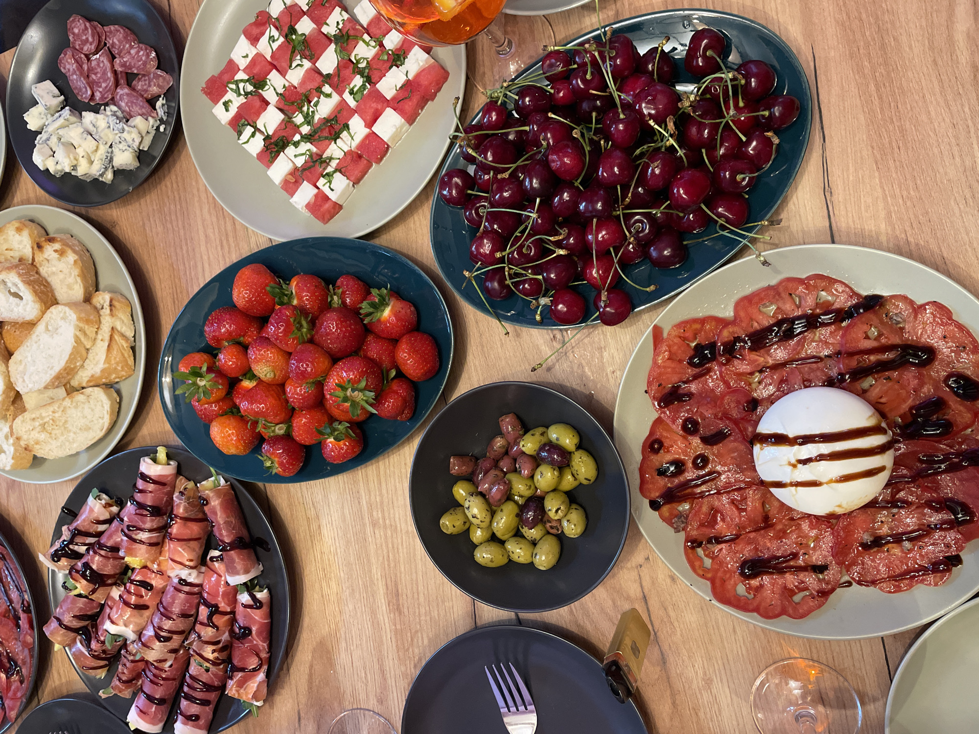 An overhead shot shows a wooden table with a variety of food items arranged in plates, likely a spread for a gathering.
In the upper left, a plate holds several prosciutto-wrapped asparagus spears drizzled with balsamic glaze. To the right of this, a plate displays sliced baguette bread. Further right, a dark plate contains slices of salami and chunks of blue cheese.
Dominating the center of the image is a dark blue plate filled with fresh strawberries. To its right, a light gray plate features a checkerboard pattern of cubed watermelon and feta cheese, garnished with chopped herbs.
Below the strawberries, another dark blue plate is overflowing with dark red cherries.
In the bottom center, a light gray plate presents thinly sliced tomatoes topped with a ball of fresh mozzarella cheese and drizzled with balsamic glaze. To the left of this, a dark gray plate holds a serving of mixed olives. A silver fork rests on this plate. To the left of the tomato and mozzarella dish, a clear glass with an orange beverage and garnish sits on the table.
The overall scene is of a well-prepared, appealing appetizer spread. The location is not discernible from the image.