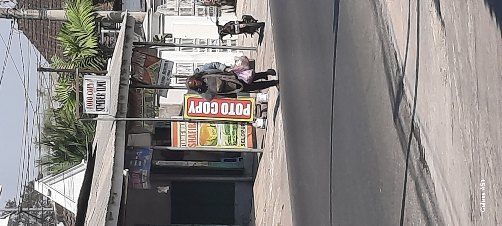 The image depicts a street scene in Selopuro, Indonesia. In the foreground, a paved road is visible. In the mid-ground, a person with dark, long hair is walking, carrying two plastic bags in their right hand. They are wearing a dark jacket over a lighter-colored garment and dark pants. To the left of the person, a stall with a sign that reads "TERANG BULAN SHAFIR SELOPUR" with an image of a food item is present. Next to it, a vertical sign with "POTO COPY" in red letters on a yellow background stands. Further to the left, a building with a covered awning and visible signage can be seen. Behind the stall, a motorcycle is parked. In the background, trees and buildings, including a house with a tiled roof, are visible under a clear sky. The overall impression is a commercial area on a sunny day.