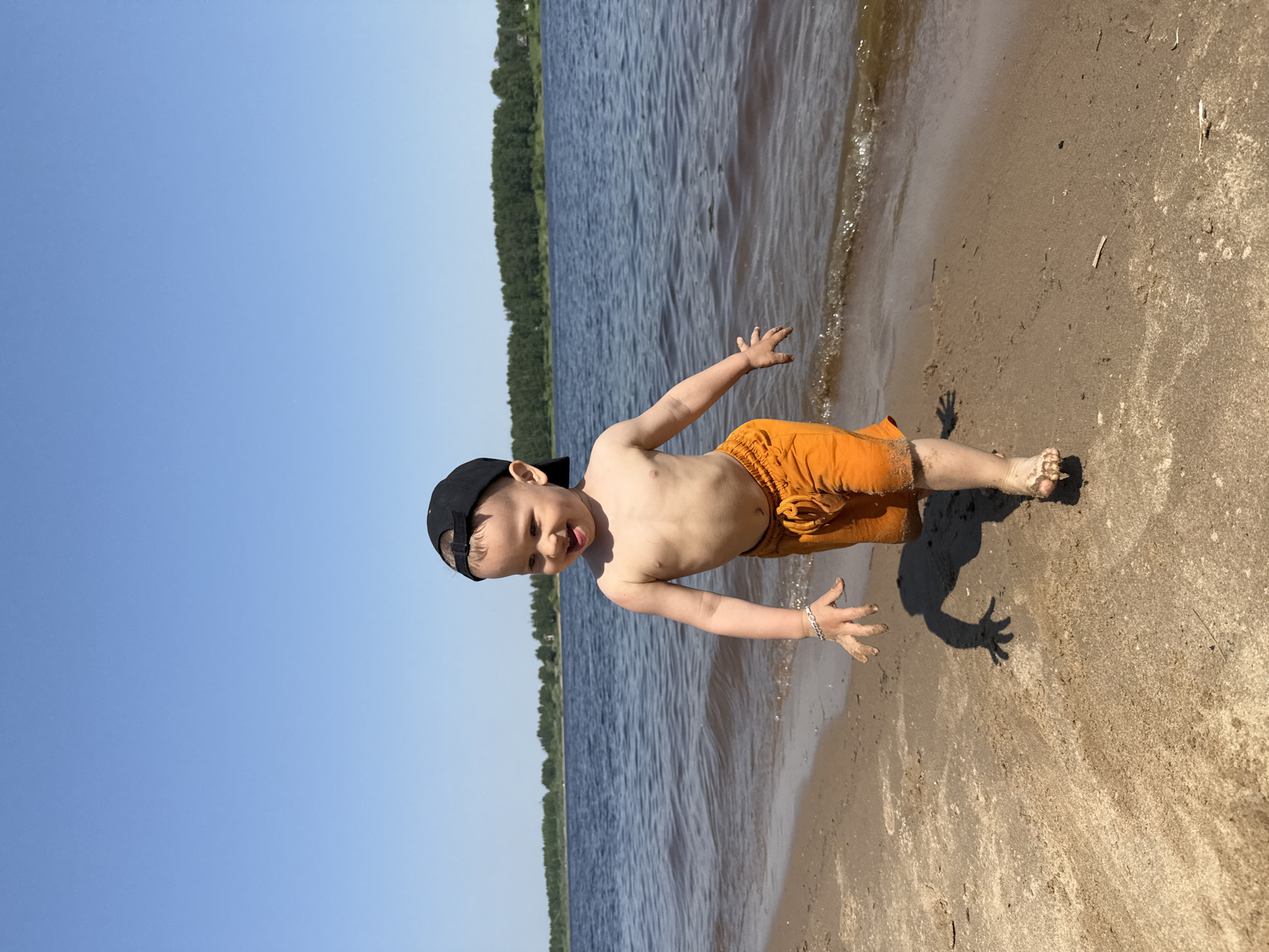 A small child wearing a black baseball cap and orange swim trunks stands on a sandy beach with arms outstretched. The child's feet and hands are sandy. Behind the child is a body of water, with a tree line visible on the opposite shore, and a blue sky. The scene is illuminated by bright daylight. There is no definitive evidence to confirm that this image was taken in the city of Rusanovo, Russia.