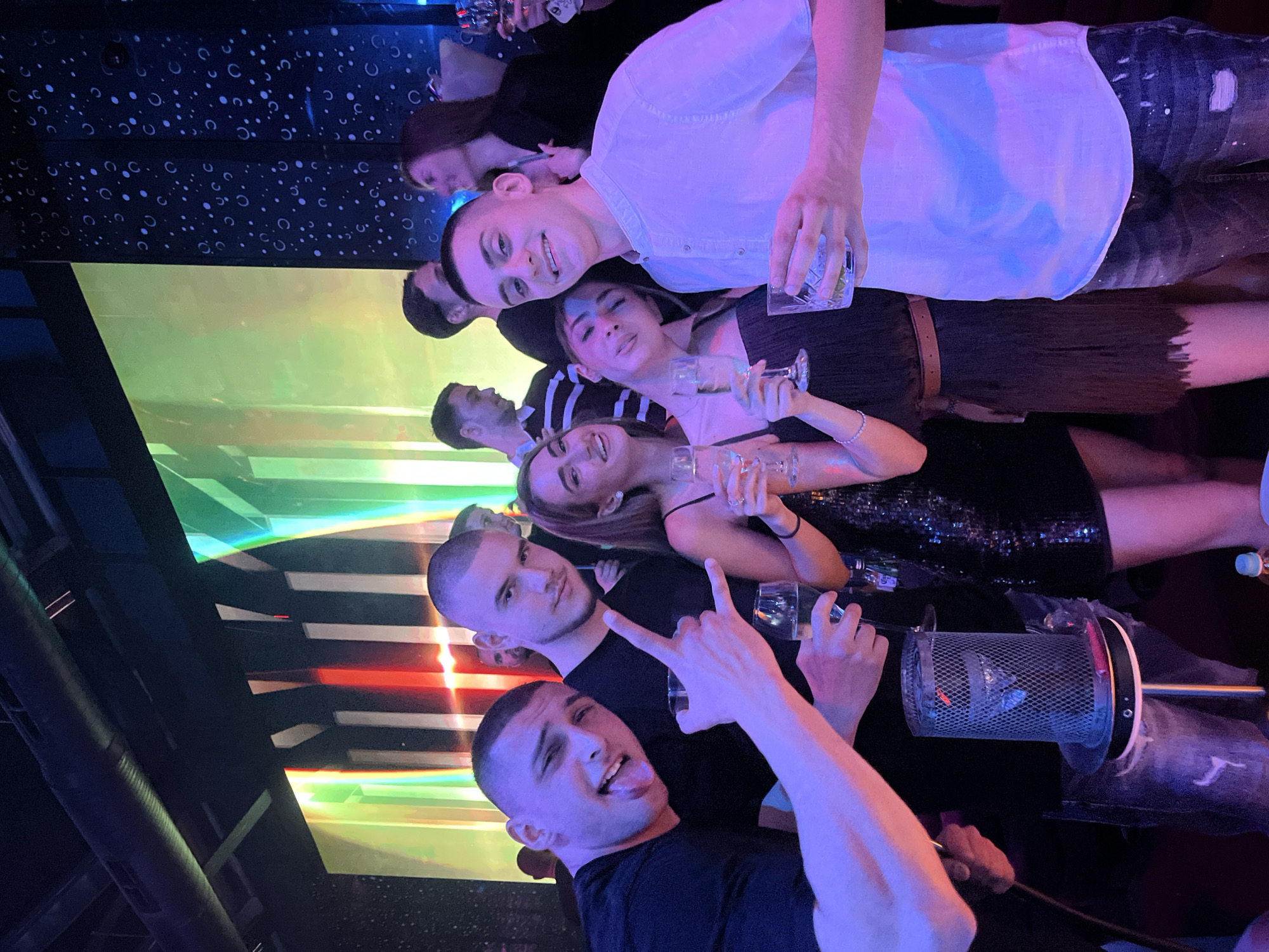 A group of approximately eight people are gathered in a dimly lit indoor setting, possibly a nightclub or bar. They appear to be posing for a photograph. Many are holding glasses, and there is a hookah present in the foreground. The location is likely Burgas, Bulgaria, as the photograph appears to be related to this city. The background features a patterned wall and colorful lighting elements. The people are dressed in a mix of casual and dressy attire.