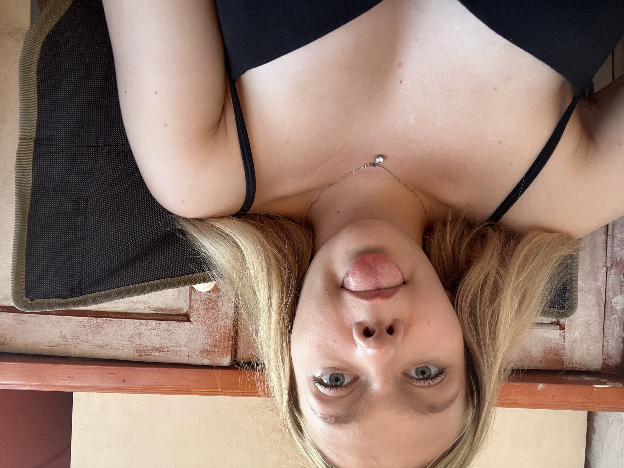 A young woman with blonde hair and blue eyes is pictured sitting in a chair, sticking her tongue out at the camera. She is wearing a black tank top and a silver necklace with a small pendant. The background appears to be indoors, with wooden paneling and furniture visible. The image does not contain any information to suggest the location is in the city of Perm, Russia.