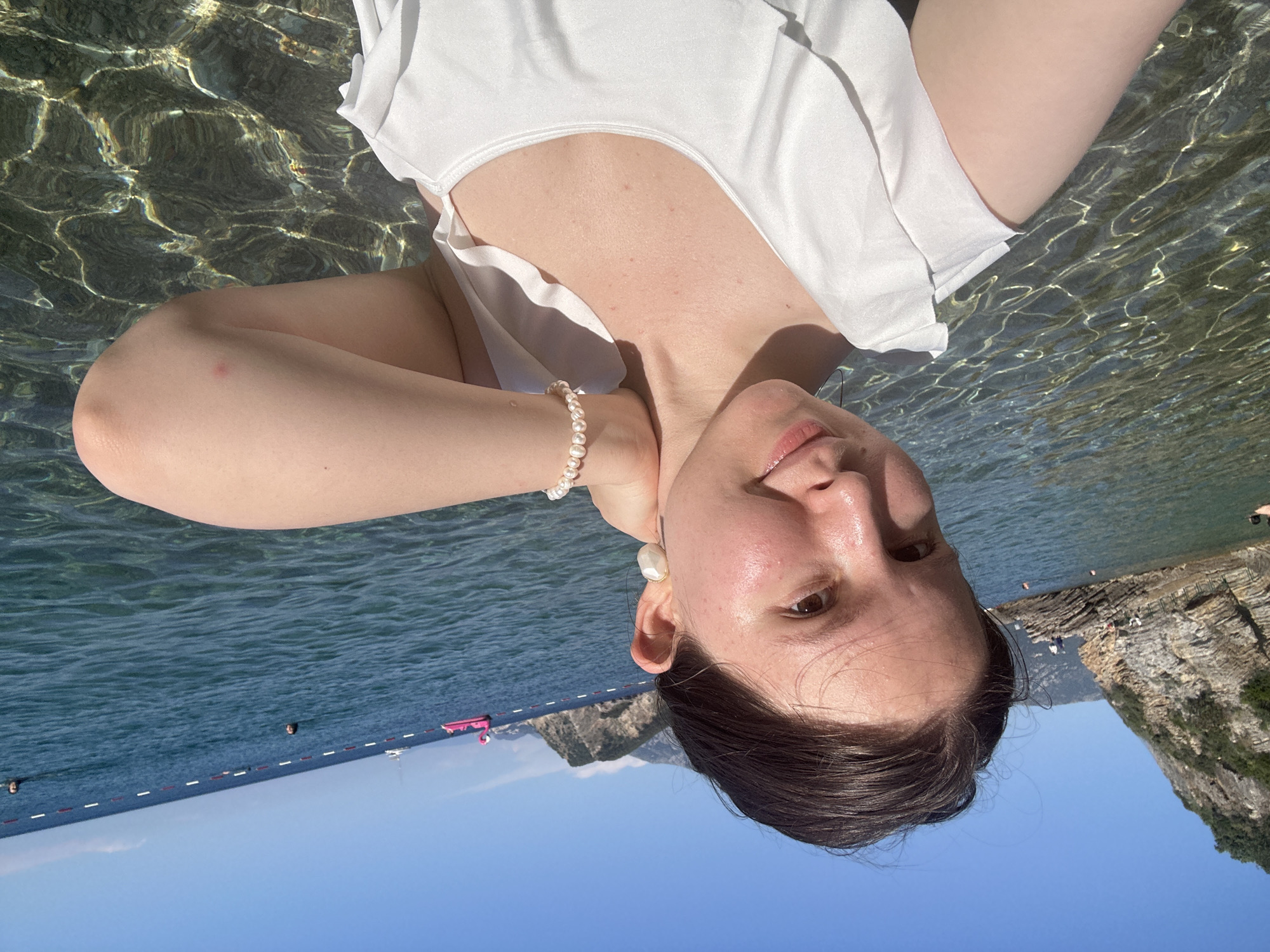 A young woman in a white top poses in the sea at Mogren Beach, Budva, Montenegro. The camera is positioned low and at an angle, partially submerged, showing the woman from the chest up. The water is clear and turquoise, reflecting sunlight and creating rippling patterns. The woman wears a pearl bracelet and stud earring. Behind her are coastal cliffs and a blue sky. In the distance, a rope barrier and buoys mark off a swimming area.