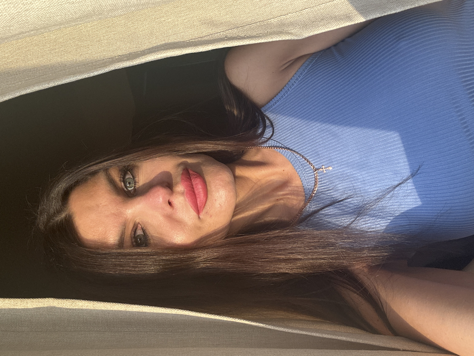 This media file is a portrait of a woman. She is lying down, with her head tilted, and her eyes are looking directly at the camera. She has long, dark brown hair, and her makeup includes bright pink lipstick and eyeliner. She is wearing a blue, ribbed tank top, and a delicate rose gold necklace with a small cross pendant. The lighting suggests it is daytime, with a warm glow casting shadows on her face and shoulder. The background is dimly lit and obscured, with a textured, light-colored fabric framing the top and bottom of the image. It appears the woman is lying on a surface covered with a similar textured fabric. The overall mood is intimate and serene, possibly captured during a relaxed moment. The scene does not contain any visible text or obvious location cues beyond what might be inferred from the indoor setting.