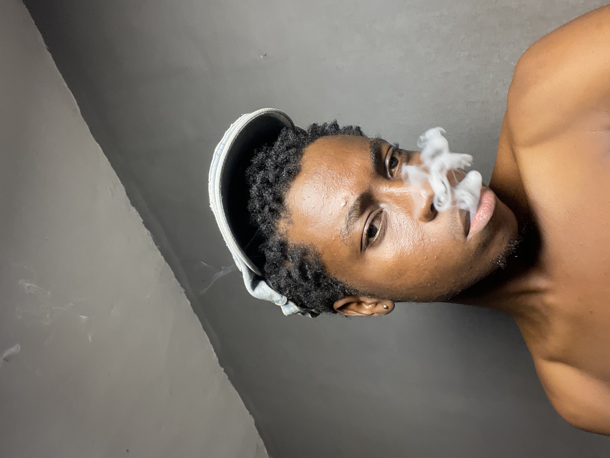 A close-up portrait, taken from a slightly low and tilted angle, captures a young man with dark skin, short, dark, curly hair, and a small gold stud earring in his left ear. He wears a light-colored cap, possibly made of denim or canvas, turned backward on his head. The man is seen exhaling a thick plume of white, wispy smoke from his mouth and nostrils, suggesting he is smoking or vaping.
He looks directly into the camera with an intense, focused gaze, his eyes being a prominent feature. The lighting highlights the contours of his face and imparts a sheen to his skin. Part of his bare shoulder and upper chest are visible in the frame, suggesting he is shirtless or wearing minimal clothing. The background is a plain, undifferentiated gray wall with subtle tonal variations, offering no specific details about the setting beyond being indoors. There is no visible text in the image. The scene conveys a calm yet intense atmosphere.