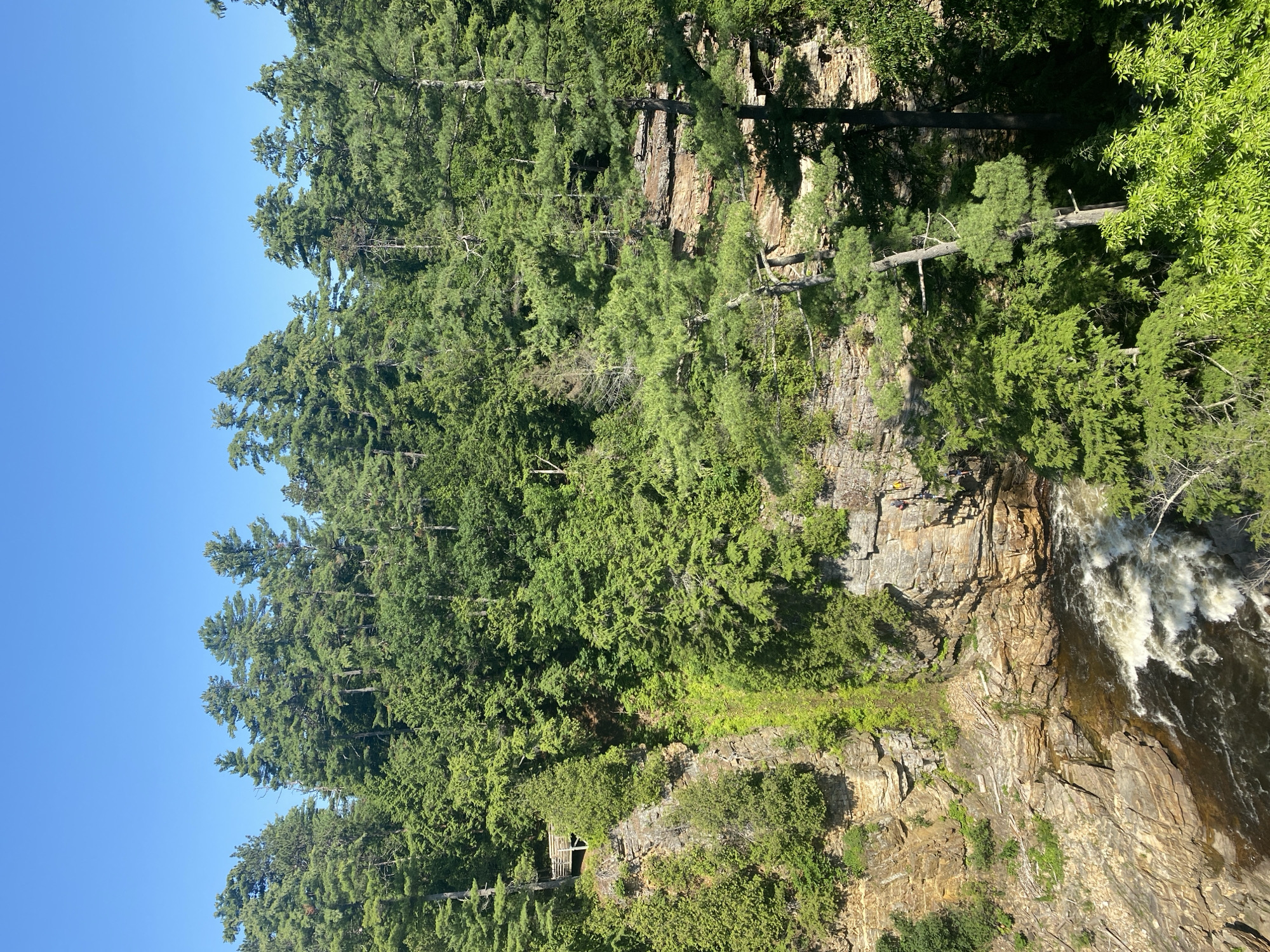 This vertical, high-angle shot captures a majestic waterfall cascading down a rocky cliff face, surrounded by a dense forest of evergreen trees. The water is a frothy white as it plunges into the dark river below. Sunlight dapples through the trees, illuminating parts of the lush green foliage and the rough, textured rock.
On the cliff face, a small wooden structure, possibly a viewing platform or a shelter, is nestled amidst the trees and rocks, adding a man-made element to the natural landscape. Several figures, appearing to be people, are visible standing on the edge of the cliff above the waterfall, seemingly enjoying the breathtaking view.
The sky is a clear, vibrant blue, suggesting a sunny day, likely in the late morning or early afternoon due to the bright lighting and clear visibility. The overall scene evokes a sense of awe and the raw beauty of nature, with the sounds of the roaring waterfall implied by the visual dynamism. No text is visible in the image. The location is the Town of Peru, USA.