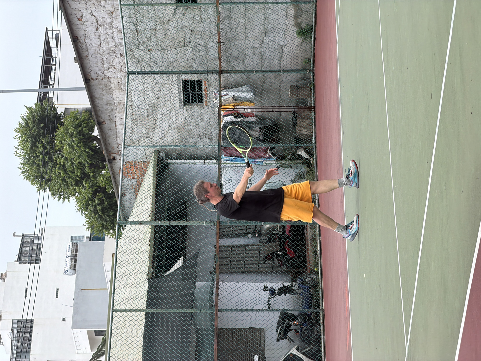 The image shows a man playing tennis on an outdoor court during the daytime. He has medium-length hair tied back and is dressed in a black t-shirt, yellow shorts, and blue and grey athletic shoes. He holds a neon yellow and black tennis racket with both hands, appearing to be in mid-action, either preparing to strike or having just struck a tennis ball.
The tennis court features a green playing surface with red boundaries and crisp white lines. A tall, dark green chain-link fence runs parallel to the court, separating it from the urban surroundings of Bac Nha Trang Ward, Vietnam. Beyond the fence, the background is densely populated with various buildings. Some are multi-story, white structures with visible air conditioning units and gridded windows, while others display rough, exposed concrete or brick walls. Green trees are interspersed among the buildings, providing natural elements.
Behind the lower section of the fence, typical urban elements include several parked motorbikes, along with various bags and other stored items. The bright, even illumination suggests a clear or possibly overcast daytime sky. The man appears focused and engaged in his game, set against the backdrop of a vibrant Vietnamese urban environment. No legible text is visible.
