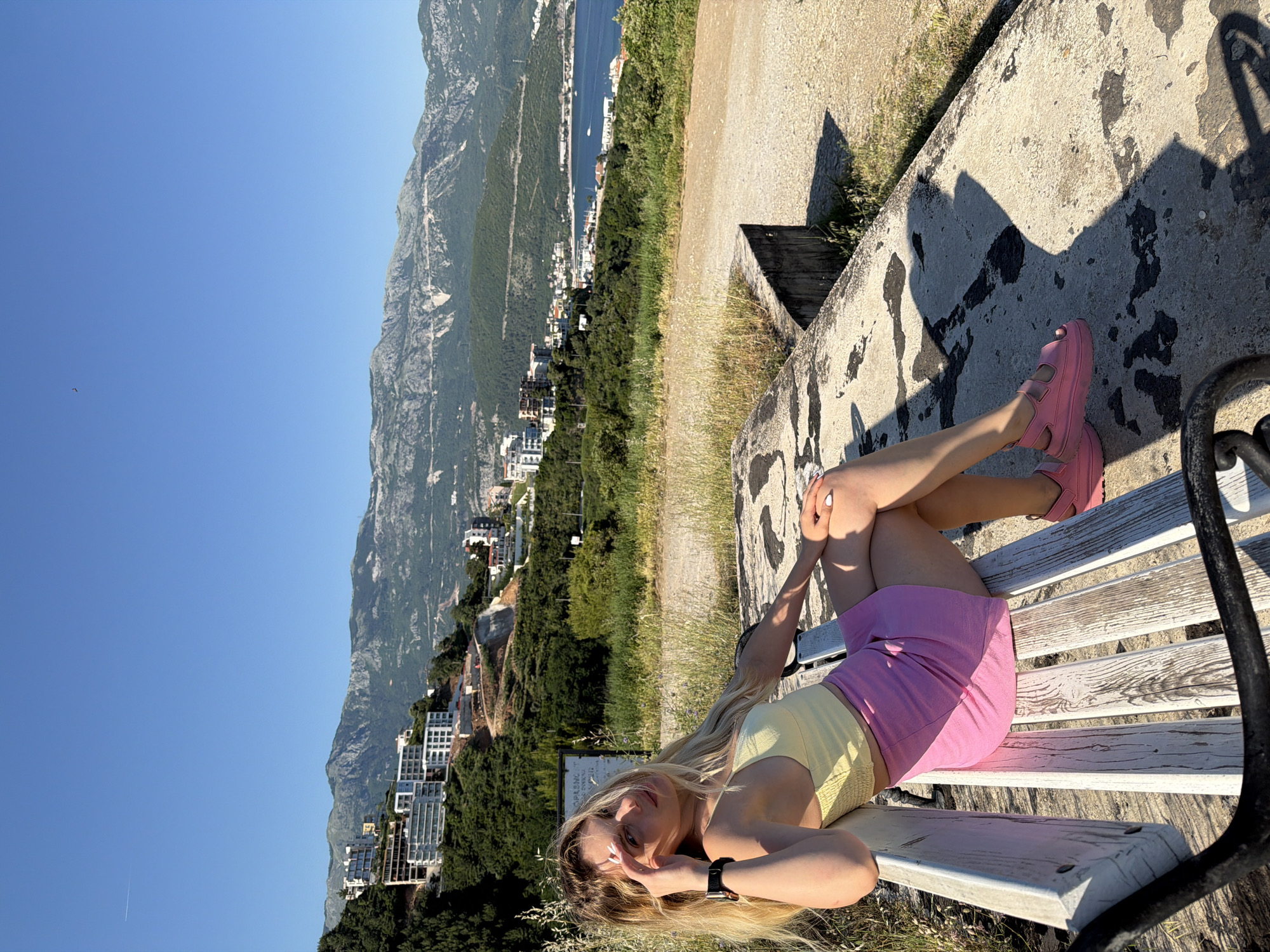 A young woman is reclining on a white bench in an outdoor location overlooking the city of Budva, Montenegro. She is wearing a yellow top, a pink skirt, and pink sandals. Her legs are crossed at the ankles, and one hand is near her face. Behind her, the city of Budva is visible, with buildings nestled between green hills and the blue Adriatic Sea. The sky is clear and blue. The bench is next to a concrete wall with dark stains and some dried grass at the base. The bench appears to be made of weathered wood and has a black metal armrest on the right. A dirt path runs behind the wall.