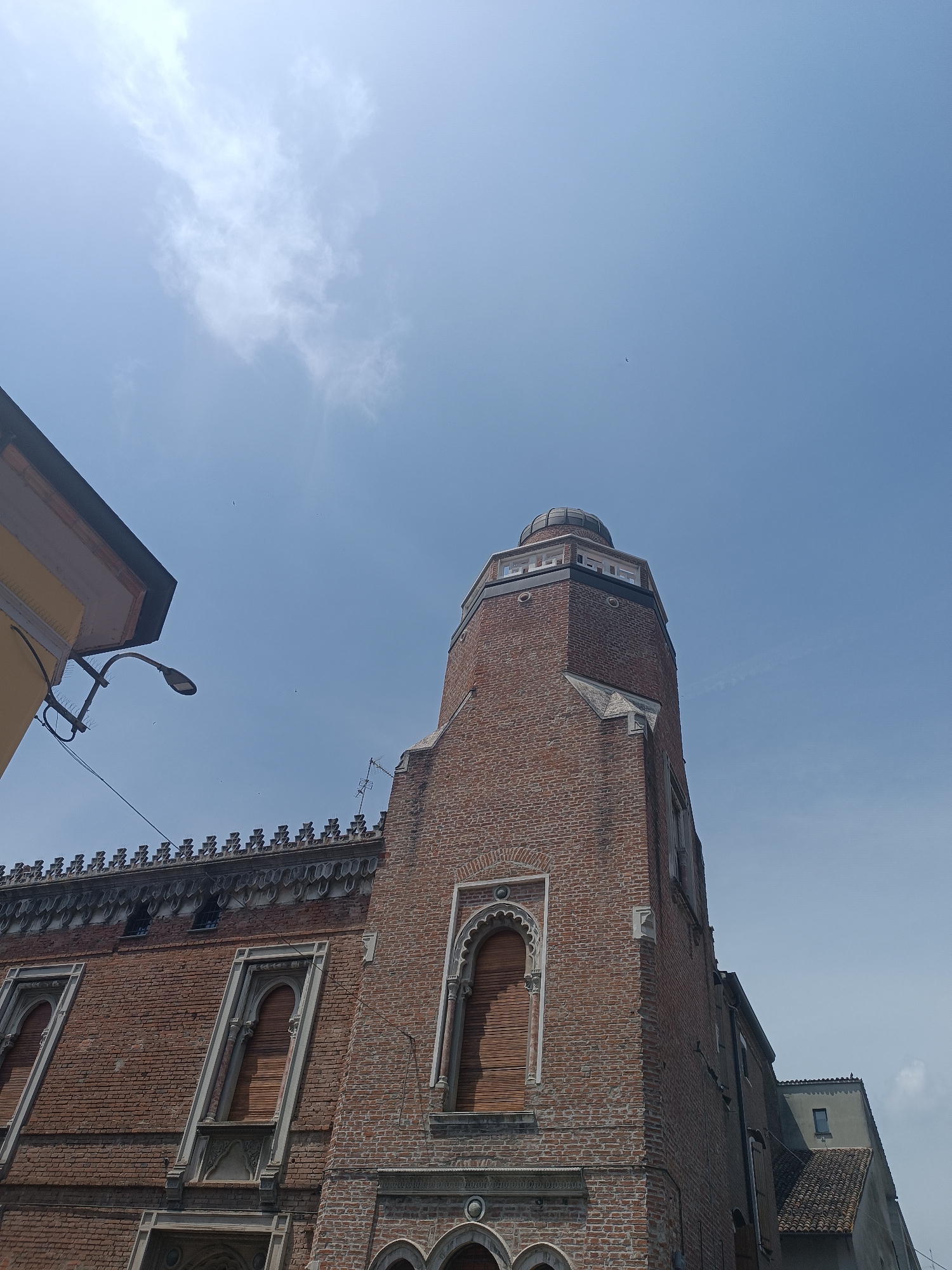 This is an eye-level, medium shot of an old brick building with a prominent tower in Casalmaggiore, Italy. The building is made of weathered red brick with arched windows featuring decorative stonework. The tower is tall and narrow, also made of brick, and topped with a small dome. The sky above is a clear blue with some wispy white clouds. A street lamp is visible on the left side of the image. The building appears to be unoccupied.
