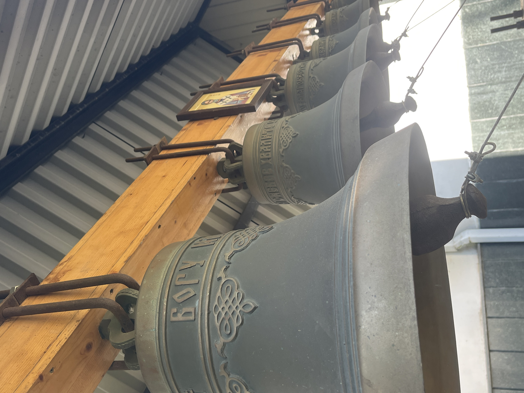 The image presents a detailed, low-angle view of multiple large bronze church bells suspended from a robust wooden beam. The foreground features the largest bell, displaying raised Cyrillic script around its upper rim, clearly spelling "БОГУ" (to God), and an ornate, intertwined decorative band below. Further along the row, subsequent bells also exhibit fainter Cyrillic inscriptions and ornamental patterns, with visible elements resembling "БЛАГОВЕСТ" (Annunciation/Good Tidings) and "ХВАЛИТЕ" (Praise). Each bell contains a dark clapper, to which thin ropes or wires are attached, extending downwards out of the frame, indicating a rigging system for ringing.
The bells are secured to the substantial, light-brown wooden beam by thick, U-shaped iron rods, which show significant rust. An Orthodox icon, framed and depicting a robed saint with a halo in traditional red and gold pigments, is affixed to the wooden beam between the second and third bells from the left. Above the wooden structure, a corrugated grey metal ceiling with distinct parallel ridges is visible, crossed by a darker horizontal beam in the distance. The scene is illuminated from the lower right, casting bright light on parts of the bells and the background. The composition and elements are consistent with the bell tower or covered entryway of a Russian Orthodox church in Krasnodar, Russia.