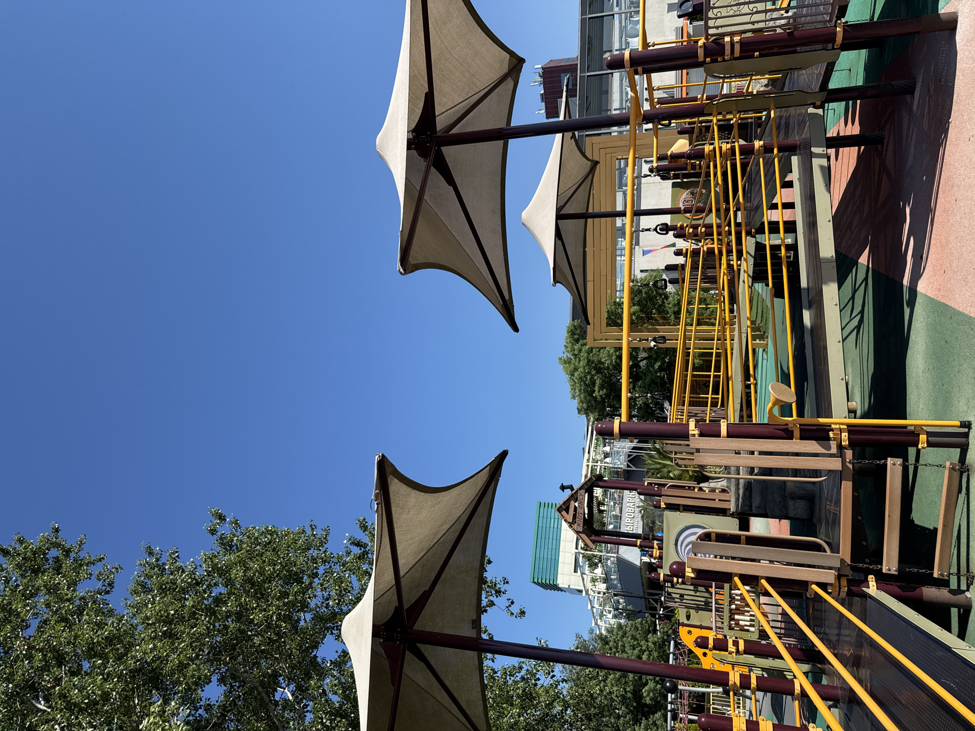 This is a ground-level outdoor shot of a modern playground on a sunny day in Sochi, Russia. The sky is a vibrant, clear blue. Two large, tan fabric shade structures with dark poles dominate the upper portion of the image. In the background, a multi-story building with a turquoise and white facade is visible, along with other structures that appear to be part of the same complex, possibly a resort or entertainment venue.
The playground features a network of yellow metal railings and maroon poles, indicating various levels and pathways. Some wooden elements, such as steps and platforms, are also incorporated into the play structure. The ground is covered in a colorful rubberized surface, with sections of green, pink, and possibly red. Trees with lush green foliage are present on the left side of the frame. A glimpse of a person, wearing a light-colored top, can be seen on the playground in the middle ground. The overall impression is a well-maintained and inviting recreational space.
