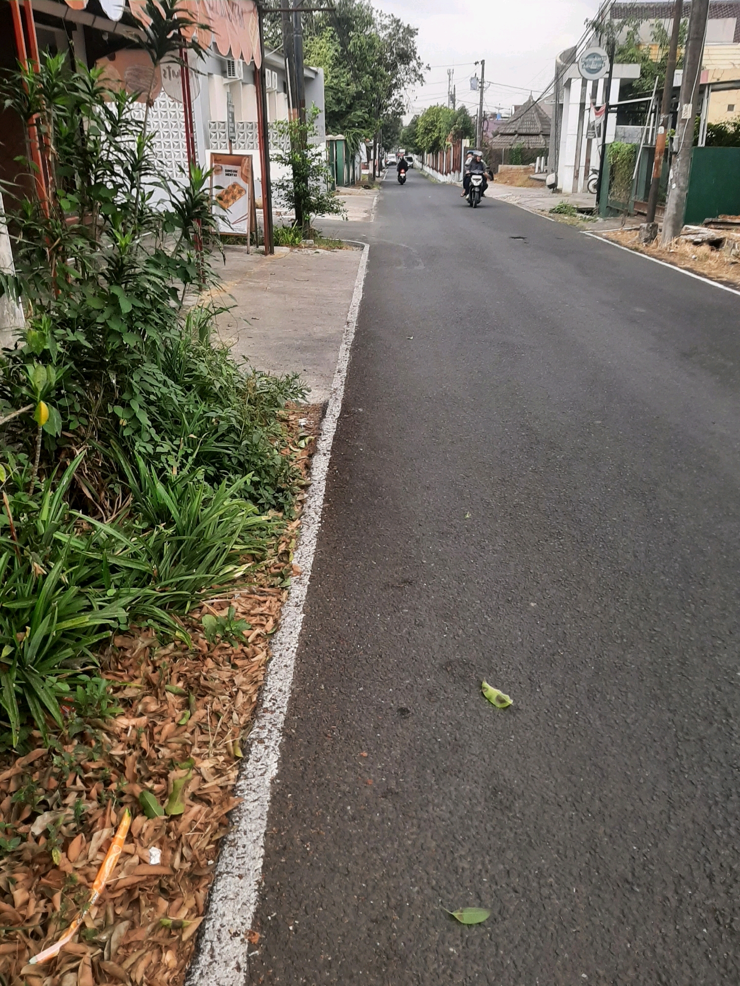 In Sumampir, Indonesia, a street scene unfolds with a paved road bordered by a concrete sidewalk and a strip of overgrown vegetation. On the sidewalk, a stand displays an advertisement for food, with a menu of dishes. Several people are visible riding motorcycles on the road. A white line marks the edge of the asphalt. The roadside is littered with fallen leaves and some debris, including a long orange object. The sky is overcast, suggesting a cloudy day.