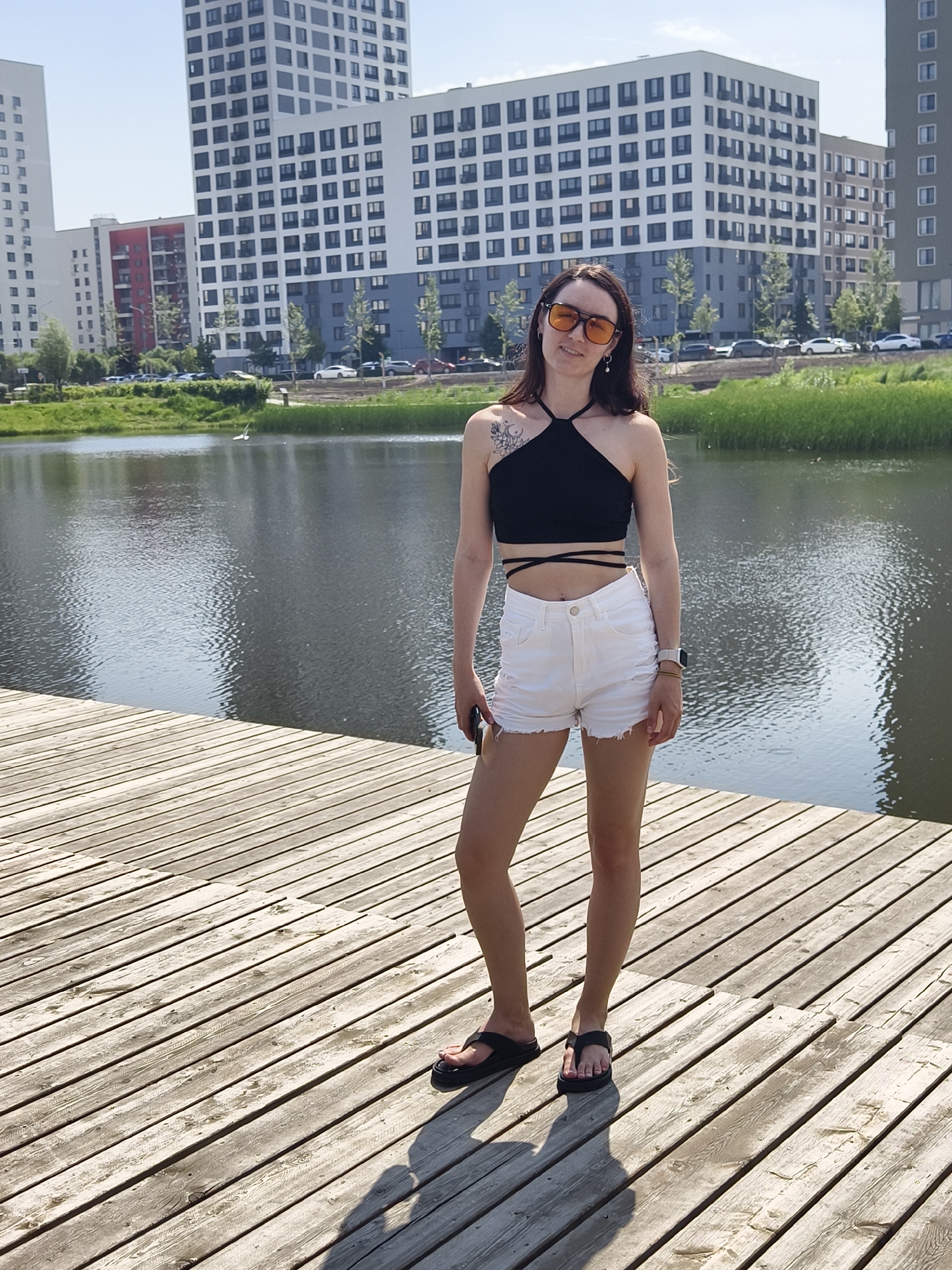 A full-body, eye-level, outdoor shot shows a young woman with long brown hair, wearing a black halter crop top, white ripped denim shorts, and black flip-flops, standing on a wooden pier. She is looking towards the camera and smiling, wearing orange-tinted sunglasses and a pearl earring. A tattoo of a crescent moon and stars is visible on her upper left shoulder. She is holding a smartphone in her right hand.
Behind her is a body of water, reflecting the sky and surrounding buildings. The buildings are modern, multi-story residential complexes, with a mix of white, blue, and red facades. Trees and grass line the edge of the water. The wooden pier is made of light-colored planks, with visible grain and some water stains. Shadows from the woman and the pier are cast on the wood. The sky is clear and blue, indicating a sunny day. The environment suggests a residential area with a water feature, possibly a park or a waterfront development in Tyumen, Russia.