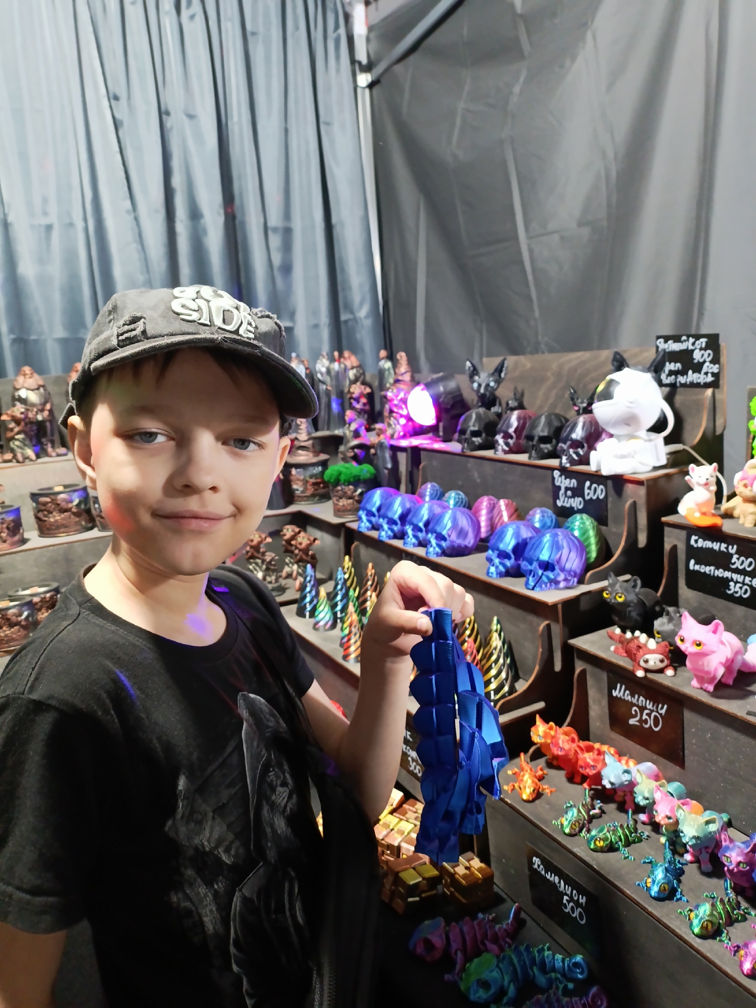 A young boy wearing a black t-shirt and a black baseball cap is standing in front of a display of 3D-printed figures. He is holding a blue, articulated sculpture in his right hand. The display features various figures including skulls, cats, lizards, and other fantasy creatures, some of which have price tags written in Russian. The background consists of grey curtains. This scene appears to be at a market or convention, possibly in Lagunovo, Russia, given the presence of Russian text.