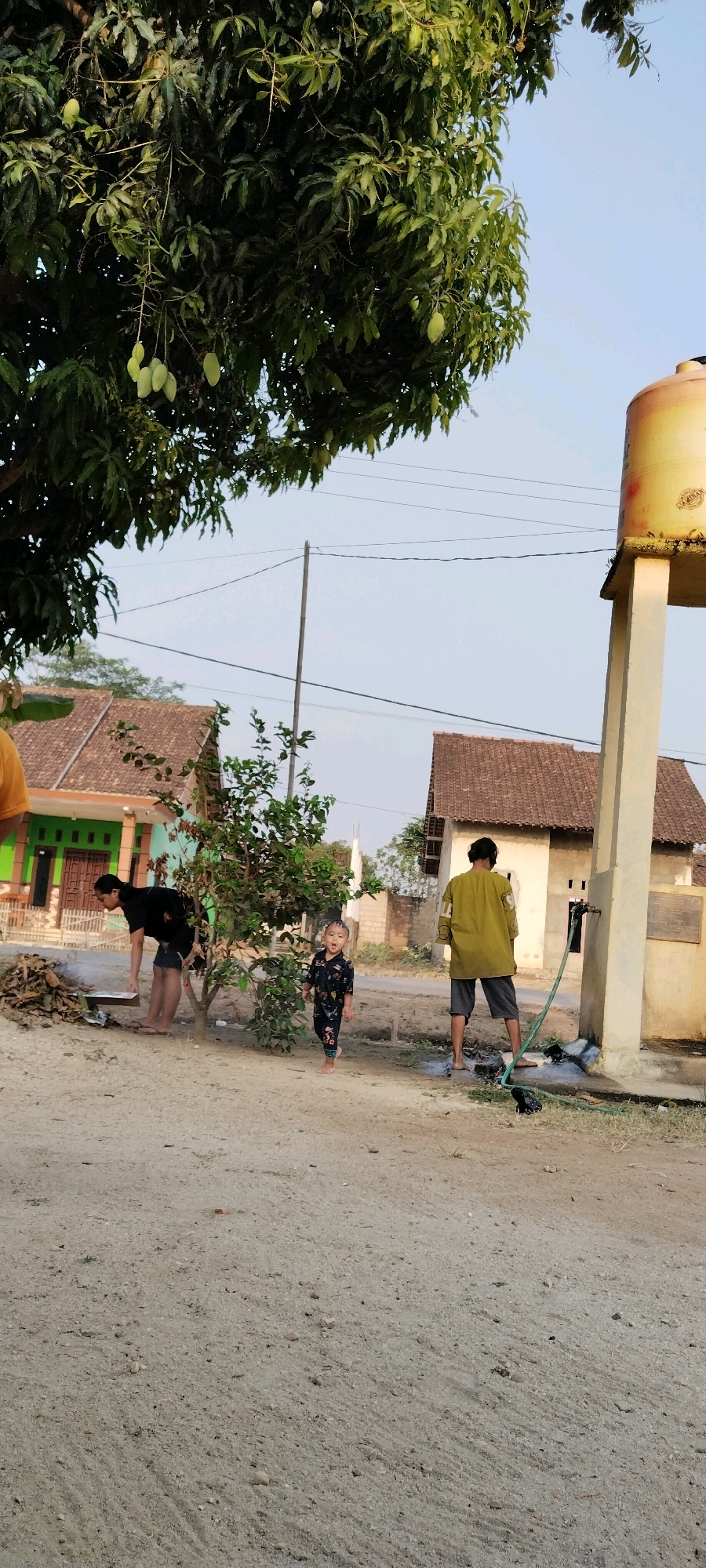 In Tanjung Ratu Ilir, Indonesia, a scene unfolds on a dirt road. A man in a black t-shirt and shorts is bending down near a pile of firewood, appearing to be engaged in an activity. To his right, a young child in a patterned outfit is standing and looking towards the camera with an open mouth, seemingly surprised or calling out. Further to the right, an individual wearing a yellow long-sleeved shirt and dark shorts stands by a water tower with a yellow tank, holding a green hose connected to a faucet. Water is visible on the ground around the faucet. In the background, residential buildings with tiled roofs are present, along with a mature mango tree laden with green fruit. The sky is clear and blue, indicating a daytime setting. The overall environment suggests a rural or semi-rural residential area.