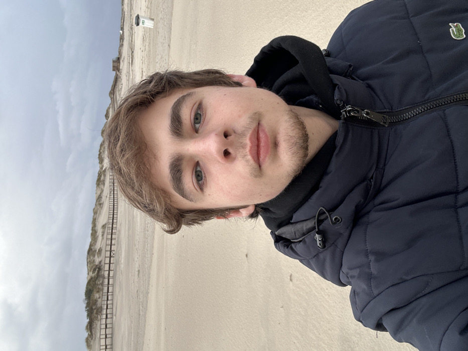 A young, light-skinned male with light brown hair, green/hazel eyes, and light facial stubble occupies the foreground, looking directly at the camera. He wears a navy blue puffer jacket with a black zipper, over a black hooded sweatshirt. A small green crocodile logo is visible on the left chest of the jacket. The scene is set on a wide, sandy beach in Gafanha da Nazaré, Portugal, under an overcast sky dominated by grey clouds. In the mid-ground, a wooden boardwalk runs horizontally across the sand, leading to a series of vegetated sand dunes. To the far right, a small wooden structure or hut is visible past the dunes, and a white and grey trash receptacle stands on the sand closer to the foreground. The lighting is diffuse.
