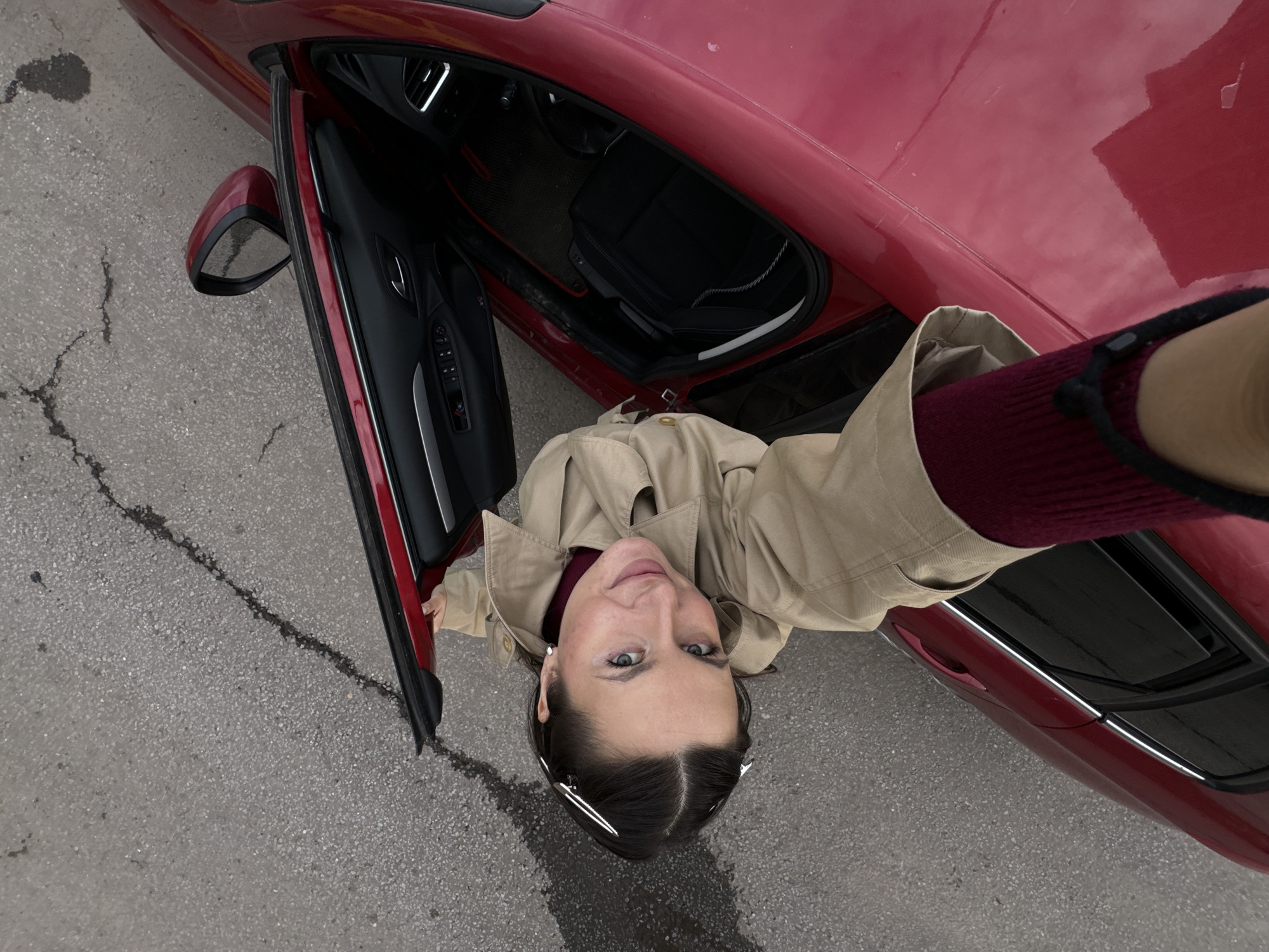 An overhead, high-angle shot captures a woman standing beside an open red car door on an asphalt surface, likely in Akulovo (Gp Kubinka), Russia. The woman, positioned centrally, looks directly up at the camera with a neutral expression and a slight smile. She has dark hair, blue eyes, and is wearing a light beige trench coat over a maroon long-sleeved top. Her right arm, holding the camera, is extended into the upper right corner of the frame.
The driver's side of a glossy red car dominates the right and upper portions of the image. Its door is open, revealing a black interior with car seats, the steering wheel partially visible, and door panel controls. The car's side mirror is also visible on the left. The ground is a textured asphalt with numerous cracks and some darker, possibly damp, patches. The reflections on the car's surface suggest an overcast or cloudy sky. No text is visible in the image.