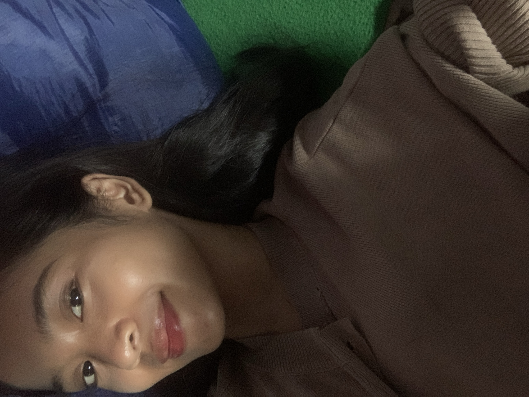 A young woman with dark hair and brown eyes is lying down. She is wearing a brown sweater. She is looking at the camera and smiling. In the background, there is a blue and green surface. The image was taken in Sungai Meriam, Indonesia.