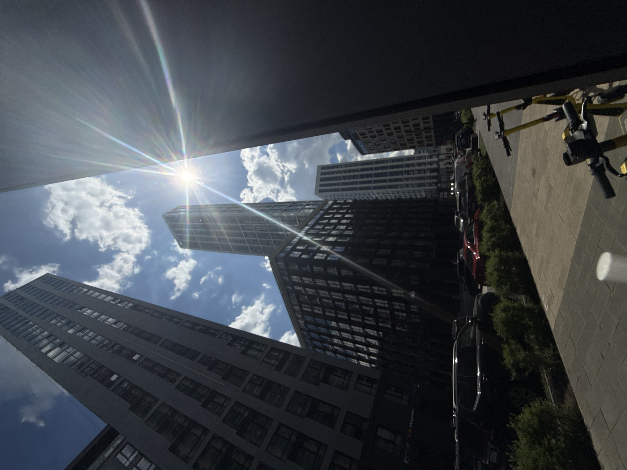 The image captures an urban landscape on a bright, sunny day, taken from a low angle looking upwards, giving a dramatic perspective on several modern high-rise buildings. The photo is oriented diagonally, with the horizon line slanted.
The sky is a clear blue with scattered, fluffy white cumulus clouds. The sun is prominently featured in the upper left, emitting intense rays and lens flare that stretch across the sky and reflect off the buildings.
Several tall, dark-colored buildings dominate the scene. Their facades are composed of numerous windows, reflecting the bright sky and clouds. One particularly prominent building stands in the center, appearing almost silhouetted against the sun. Other buildings are visible in the background and foreground, contributing to the dense urban environment.
At ground level, on the right side of the image, there is a paved sidewalk. A row of vehicles, including several parked cars (one red, one dark SUV), and a line of yellow electric scooters, are visible along the curb. Lush green bushes are planted between the sidewalk and the buildings.
Notable details include the midday sun's intensity, creating strong contrasts between brightly lit and shaded areas. The modern architectural style of the buildings suggests a contemporary city setting, consistent with Moscow. Faint text visible on a lower building's facade reads "Яндекс Маркет" (Yandex Market). There are no people visible, but the presence of vehicles and shared scooters indicates a functional urban space.