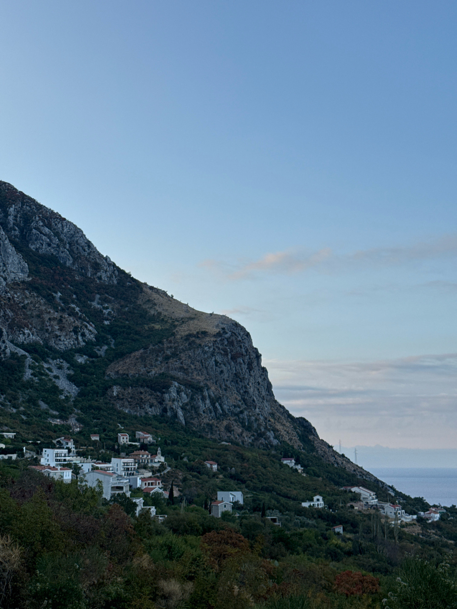 This is an outdoor, horizontal, medium-shot photograph of a coastal mountain range with a village nestled on its slopes. The sky is a clear, pale blue, with a few wispy clouds visible near the horizon. The mountain, to the left of the frame, is steep and rocky, with patches of scrubby vegetation. The village, spread across the hillside, consists of numerous white buildings with red-tiled roofs. The houses are of varying sizes and architectural styles, some appearing more modern with balconies and large windows, while others are smaller and more traditional. The village is surrounded by lush green foliage, including trees and bushes. In the distance, a body of water, presumably the Adriatic Sea, is visible beyond the lower slopes of the mountain, meeting the horizon. The lighting suggests it is either early morning or late evening. No people are visible in the image. The location is identified as Vrba, Montenegro.
