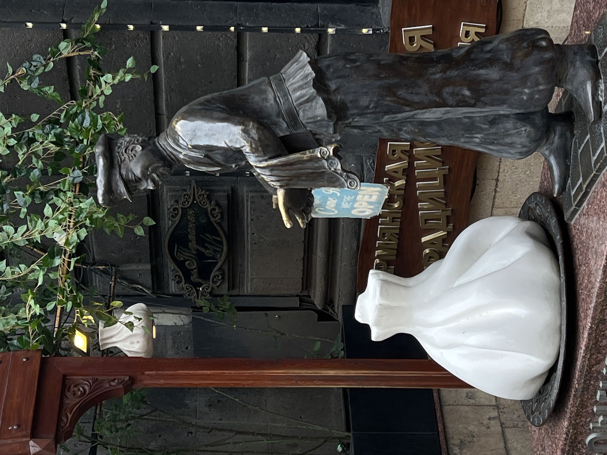 A vertical shot shows a bronze statue of a man in front of a restaurant. The man wears a long coat and hat, and he is holding a small sign that says "Come In We're Open". A large white dumpling is sitting on a plate next to him, and there is greenery on the left side of the frame. The name of the restaurant is written in Russian on a wooden sign in the background. The location is in the city of Unknown, Unknown.
