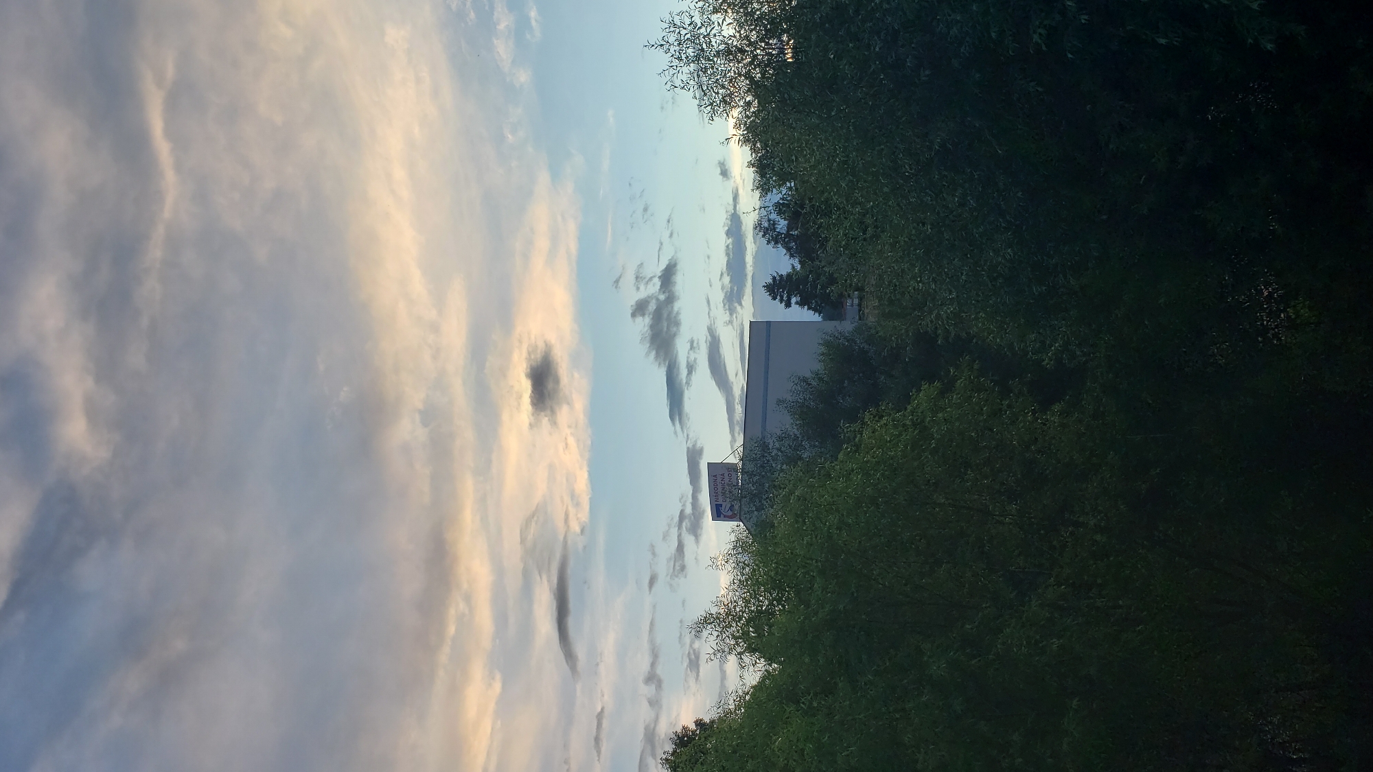 The image shows a view of the sky above a building and trees in Prešov, Slovakia. The sky is filled with clouds, some of which are illuminated by the setting sun, casting a warm glow. A white building with a sign that reads "NÁRODNÁ BANKA SLOVENSKA" is visible above a dense canopy of green trees. The building appears to be a financial institution, likely a branch of the National Bank of Slovakia. The trees are lush and full, suggesting a time of year with abundant foliage. The overall scene depicts a peaceful urban landscape bathed in the soft light of dusk.