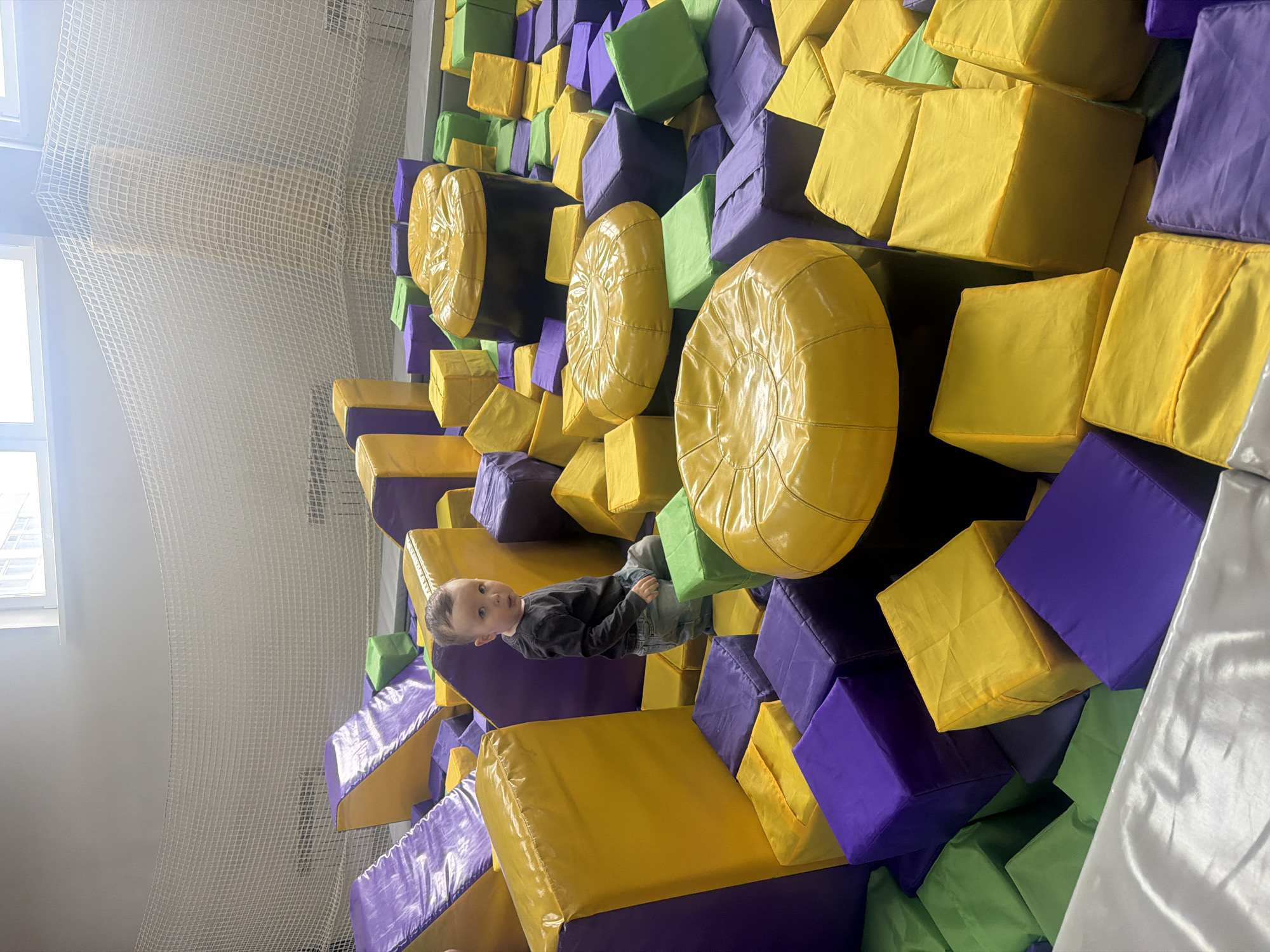 A young child is lying among yellow, purple, and green foam blocks and yellow cylindrical foam objects in what appears to be a play area, possibly an indoor playground or a foam pit. The child is wearing a dark long-sleeved shirt and denim jeans. A safety net is visible in the background on the left side of the image, near a window.
While I do not have enough information to determine the exact location within the city of Chaikovsky, Russia, the scene appears to be within a recreational or entertainment facility.