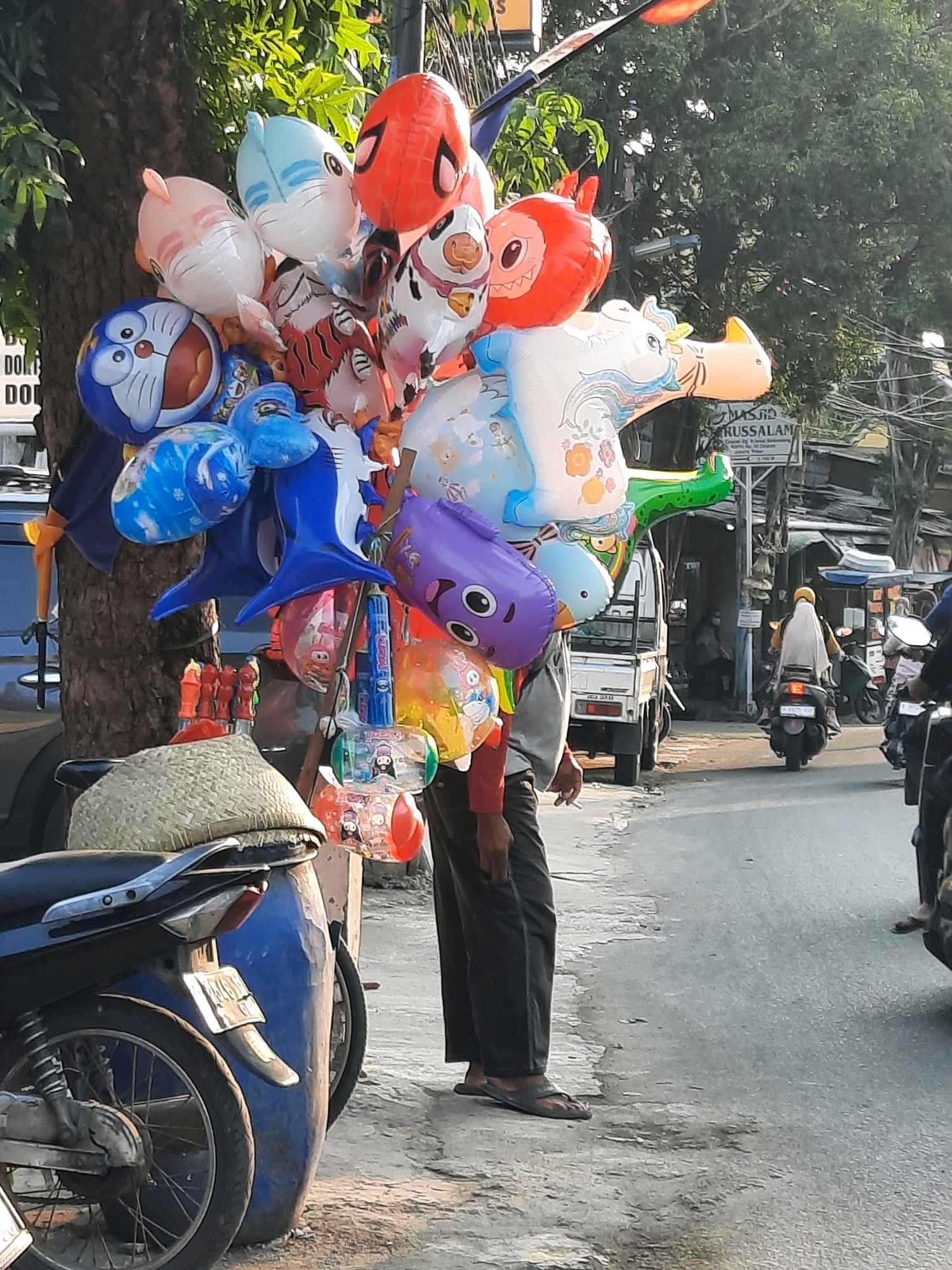 This image captures a street scene in East Jakarta, Indonesia, focusing on a vendor selling balloons on the sidewalk. The vendor, a man wearing dark pants and flip-flops, stands beside a large collection of colorful foil balloons. The balloons are attached to poles and vary in design, featuring characters like Doraemon, a shark, a tiger, and a horse, as well as a Spider-Man themed balloon. Some of the balloons are partially obscured by a large blue barrel and a woven straw hat placed on top of it.
In the background, a white pickup truck is parked on the street, and further down, a woman wearing a yellow hijab and a long white dress is riding a black motorcycle. A sign on a building in the background reads "MASJID TRUSSALAM," indicating the presence of a mosque. The street is paved and appears to be a moderately busy urban area with buildings and trees lining the road. The lighting suggests it is daytime, possibly late afternoon given the warm tones. The overall atmosphere is that of a typical street vendor scene in Indonesia.