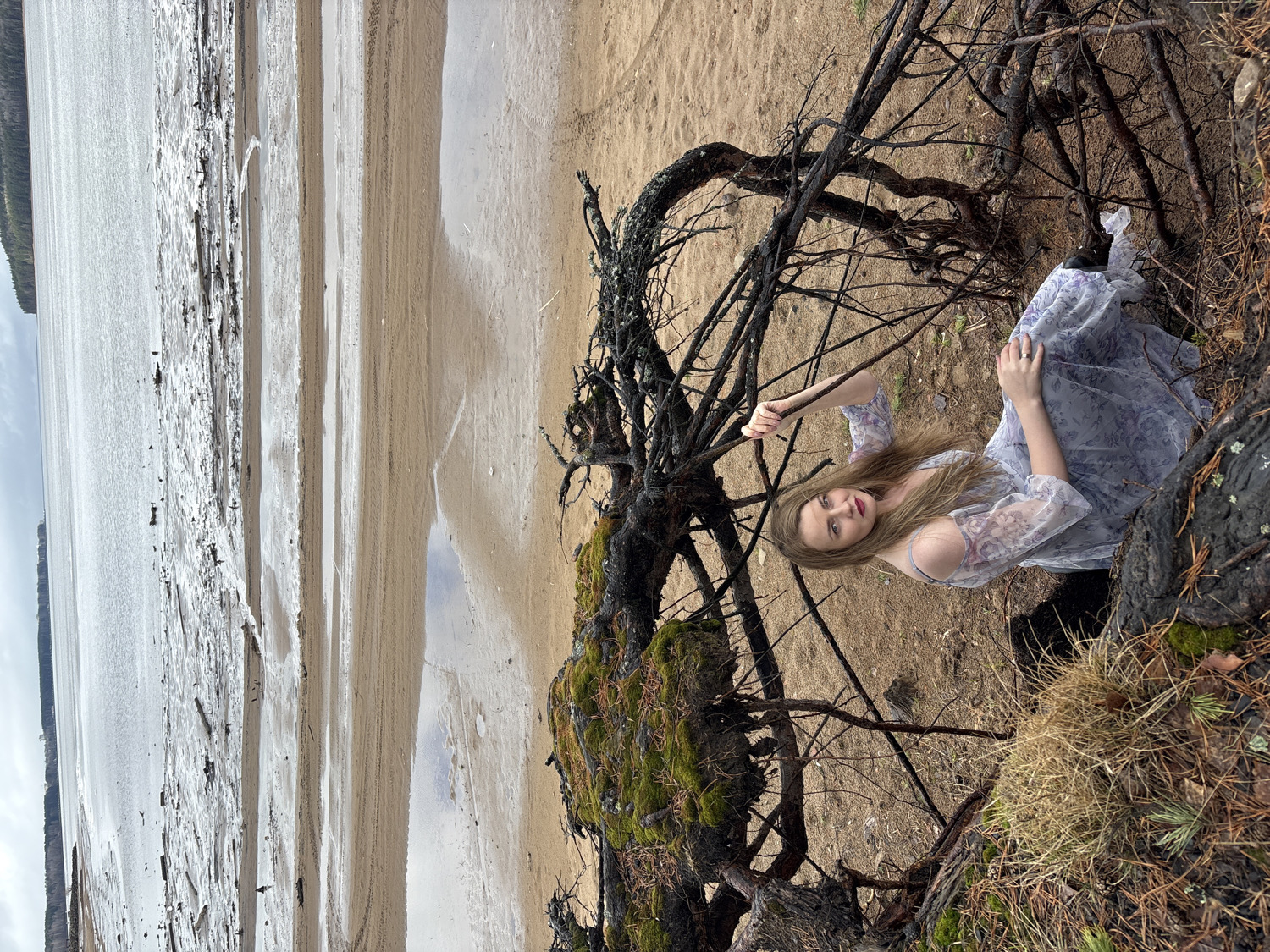 A young woman with long, light brown hair is pictured lying on a sandy bank, nestled amongst large, gnarled tree roots. She wears a light-colored, possibly light blue, long-sleeved dress with a floral pattern and looks directly towards the camera with a serious or contemplative expression. Her hands, adorned with rings, are visible; one holds onto a thin branch or root, while the other rests near her chest.
The prominent tree roots are dark and twisted, with patches of green moss growing on them, and dry grass and pine needles are scattered around them. To the left of the woman, a wide expanse of calm water, possibly a large river or lake, stretches towards the horizon. The water reflects the overcast sky, creating a silvery sheen. The sandy bank transitions into wet sand with ripples and debris, indicating fluctuating water levels. A dark, forested shoreline is visible in the distant background, beyond the water.
The scene appears to be set during daytime, likely on an overcast day given the soft, diffused lighting. The overall impression is that of a posed photoshoot in a natural outdoor environment. The location is Chaikovsky, Russia, consistent with a wide river or lake environment like the Kama River. There is no visible text in the image.