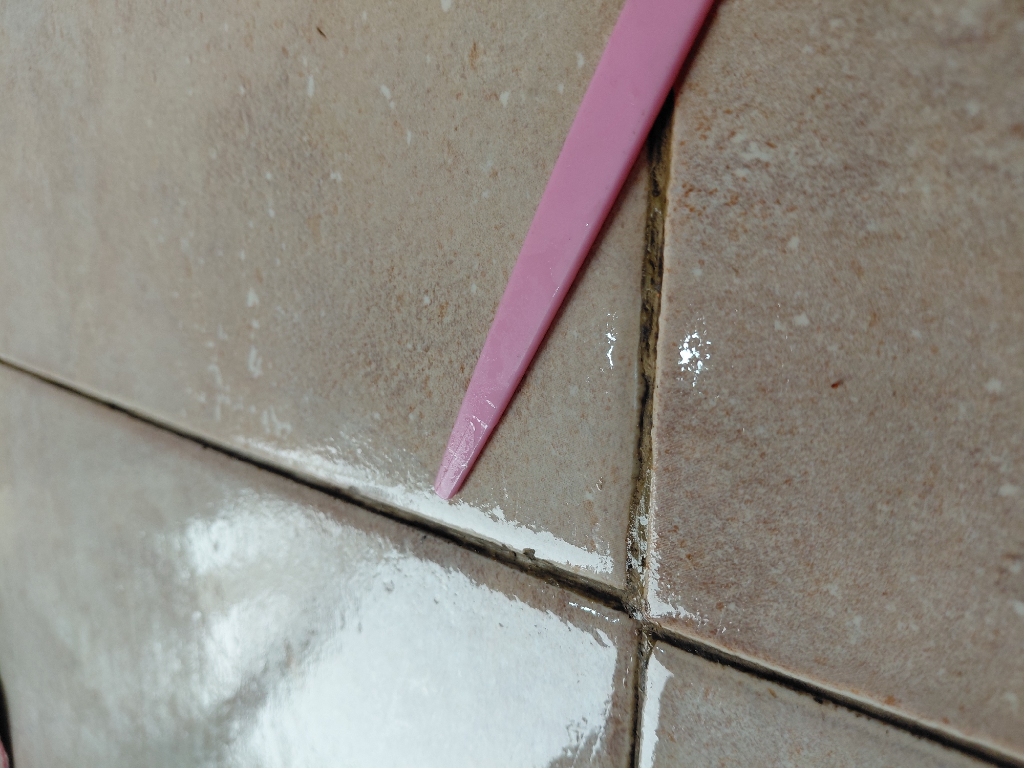 The image provides a close-up view of light brown or beige ceramic tiles, likely part of a wall or floor in an indoor setting such as a bathroom or kitchen, situated in Santa Ana, Venezuela. A pink, pointed tool, resembling a spatula or scraper, is prominently positioned in the upper center, with its tip resting on a horizontal grout line.
The tiles exhibit a slightly textured, granular surface. They appear wet or damp, with noticeable shiny reflections indicating the presence of water or a liquid film across their surfaces and within the grout lines. The grout lines themselves are dark, appearing discolored and possibly containing grime or mold in some areas. A fresh, white, pasty substance, likely new grout or caulk, is visible along the horizontal grout line where the pink tool is active, as well as splattered onto the adjacent tile surfaces.
The scene depicts an activity involving tile and grout maintenance, possibly cleaning, re-grouting, or sealing. The pink tool seems to be either applying or removing the white substance from the grout line. There are no people visible, and no text is discernible in the image. The lighting suggests an indoor environment, but the time of day and weather conditions cannot be determined.
