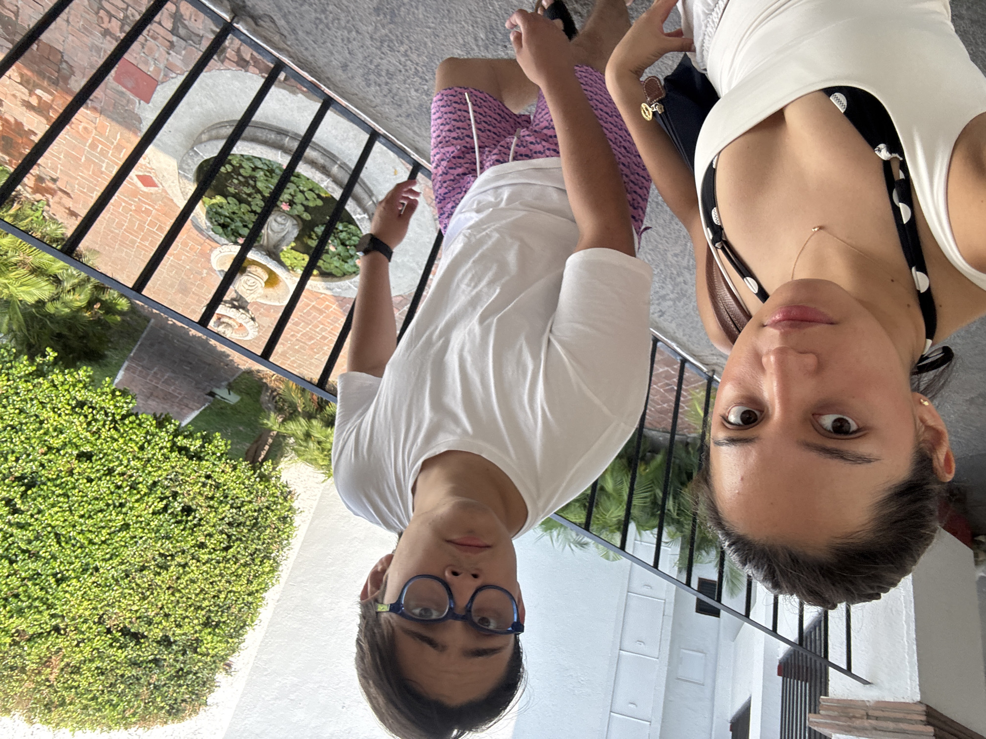 A man and a woman are posing close to each other, with the camera positioned above and angled downwards, outdoors on a balcony in Cocoyoc, Mexico. The man, wearing glasses and a white t-shirt and pink patterned shorts, is on the left. The woman, wearing a white tank top and a black and white polka dot bra, is on the right. A small water fountain is visible through a black metal railing. Lush green foliage is seen in the background.