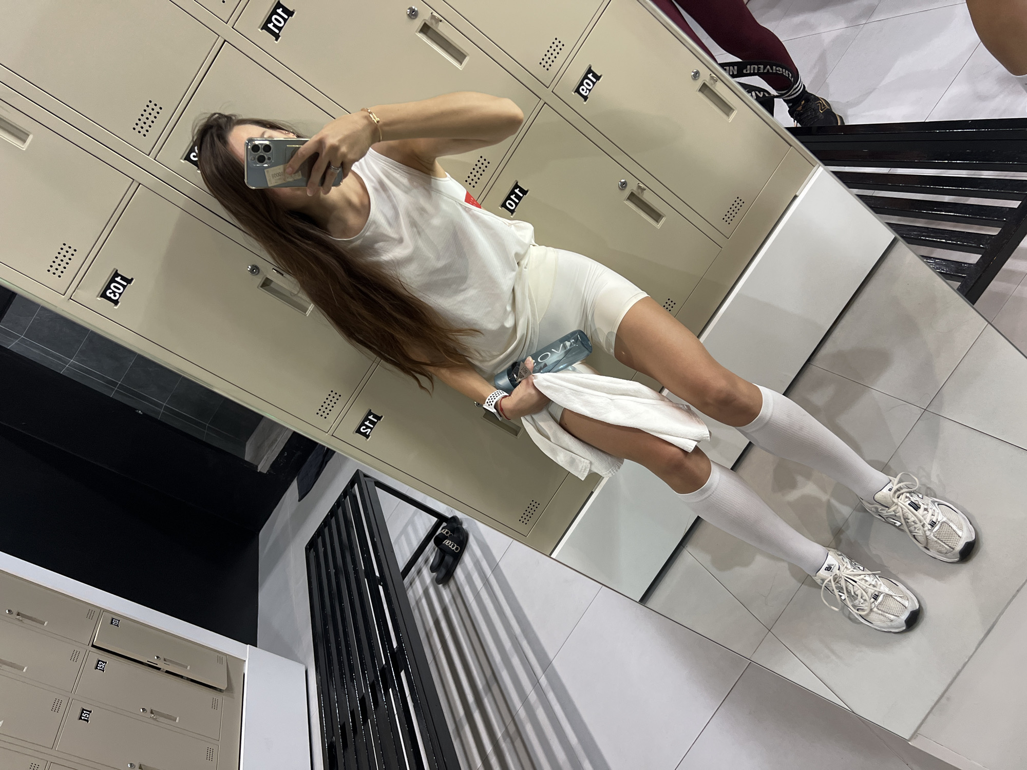 A woman is taking a selfie in front of lockers in a gym. She is wearing a white athletic outfit consisting of a tank top and shorts, along with white socks and running shoes. She's holding a phone in one hand, and a water bottle and towel in the other. The gym lockers are tan and numbered, and there is a black metal bench nearby.