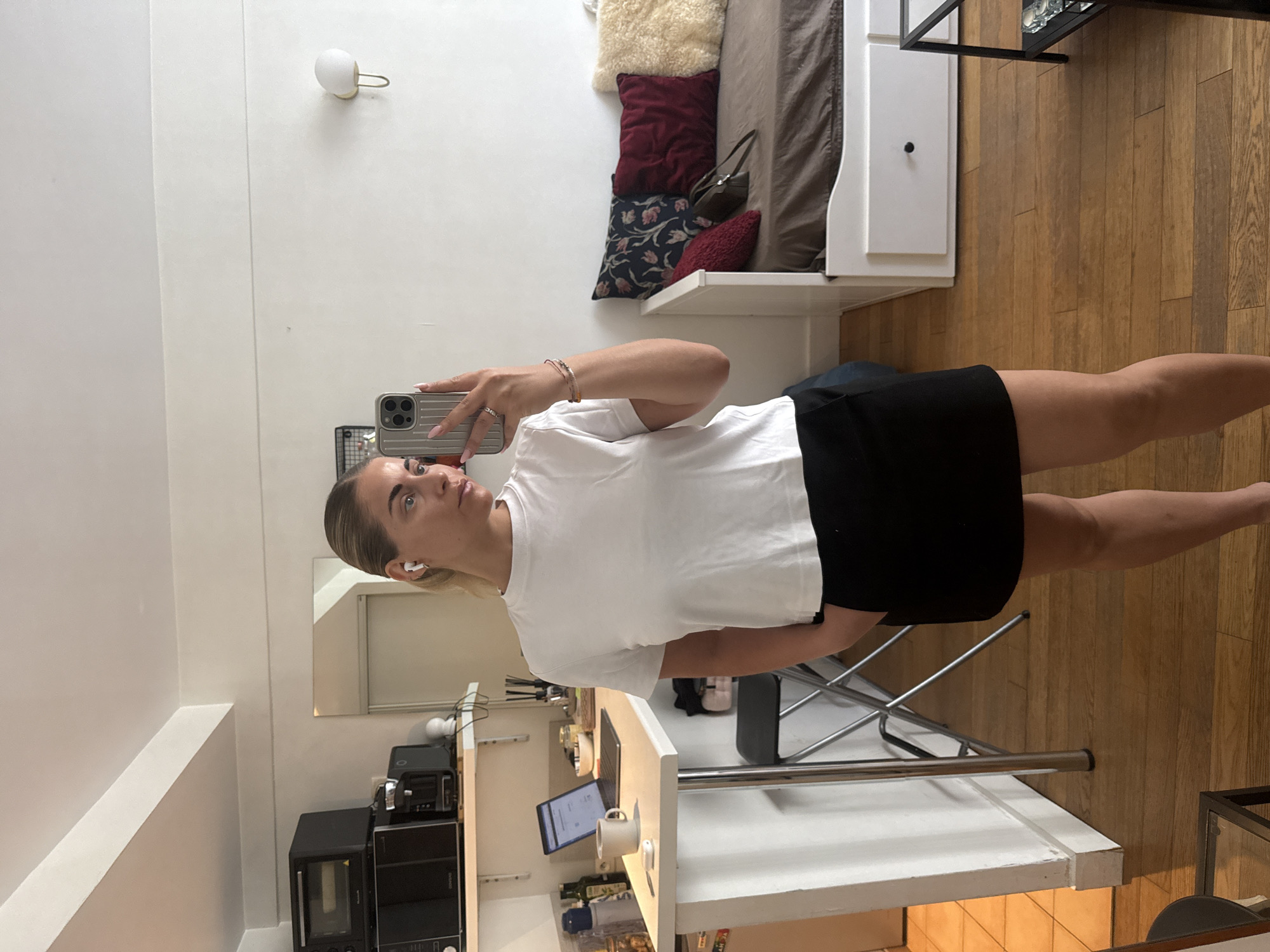 The image depicts a selfie taken in a kitchen area of an apartment, likely in Paris, France. A woman with blonde hair pulled back and wearing white earbuds is standing, holding a silver smartphone in her right hand, taking the photo. She is wearing a white short-sleeved t-shirt and a black skirt. Her left hand is in her skirt pocket. The background shows a white countertop with a laptop, a coffee mug, and various kitchen items. Further back, a microwave and coffee maker are visible on a shelf. To the right of the woman, a daybed with several pillows of different colors and textures is present. The room has wooden flooring and white walls, with a wall-mounted light fixture. The overall impression is of a compact, well-lit living space.
