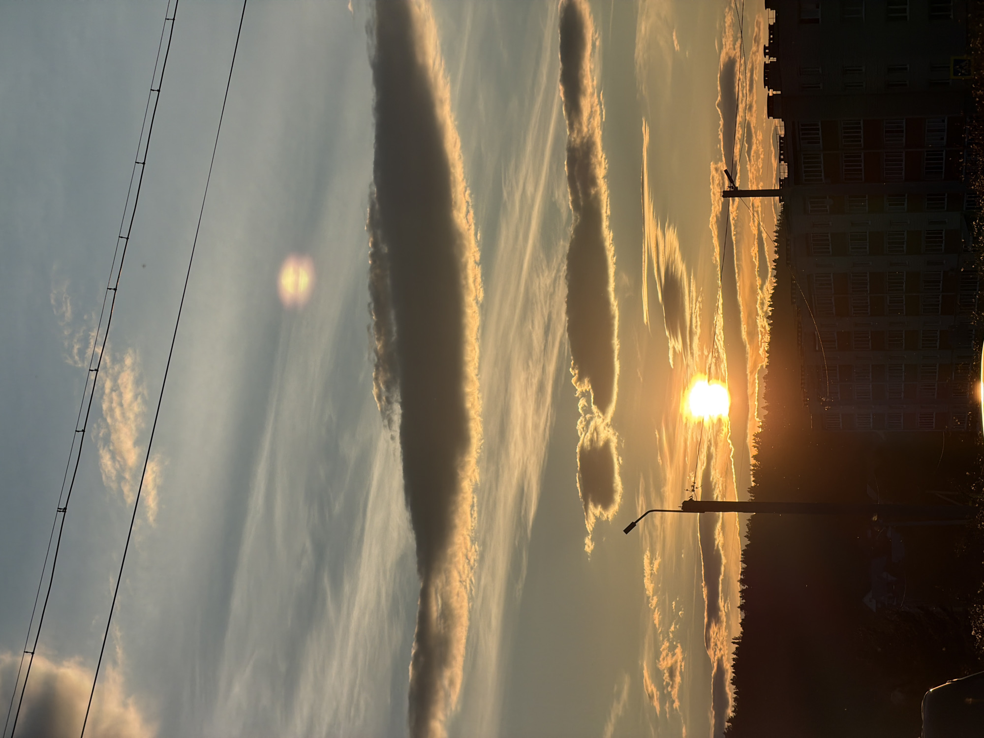 The image depicts a sunset over a city. The sun is a bright orb, partially obscured by clouds, casting a warm glow across the sky. Wispy clouds are scattered throughout, some illuminated by the sun's rays. In the foreground, silhouetted against the vibrant sky, are a series of utility poles and wires. To the right, the dark outlines of apartment buildings suggest an urban environment. A line of trees forms a dark band along the horizon, separating the sky from the buildings. The overall impression is one of a tranquil evening in an urban setting.
