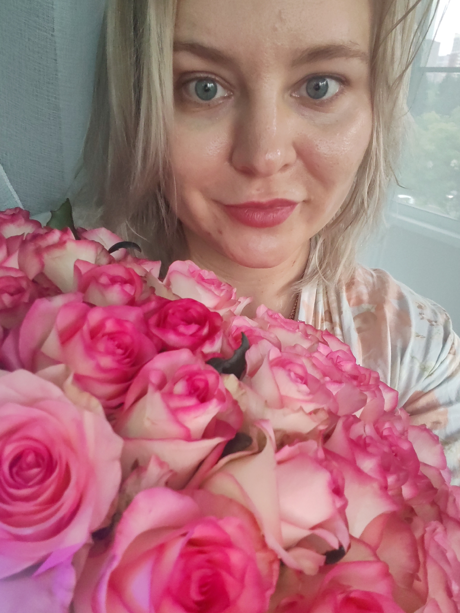 This is a close-up, portrait-oriented selfie of a Caucasian woman in Izhevsk, Russia, holding a bouquet of pink and white roses. The woman has blonde hair, blue eyes, and is wearing light-colored lipstick. She is wearing a light-colored robe with a floral pattern. The roses are in focus in the foreground, obscuring the lower part of her face and body. The background is blurred, showing a window with greenery outside, suggesting an indoor setting possibly near a window. There are no other people or significant objects visible. The lighting appears to be natural light coming from the window.