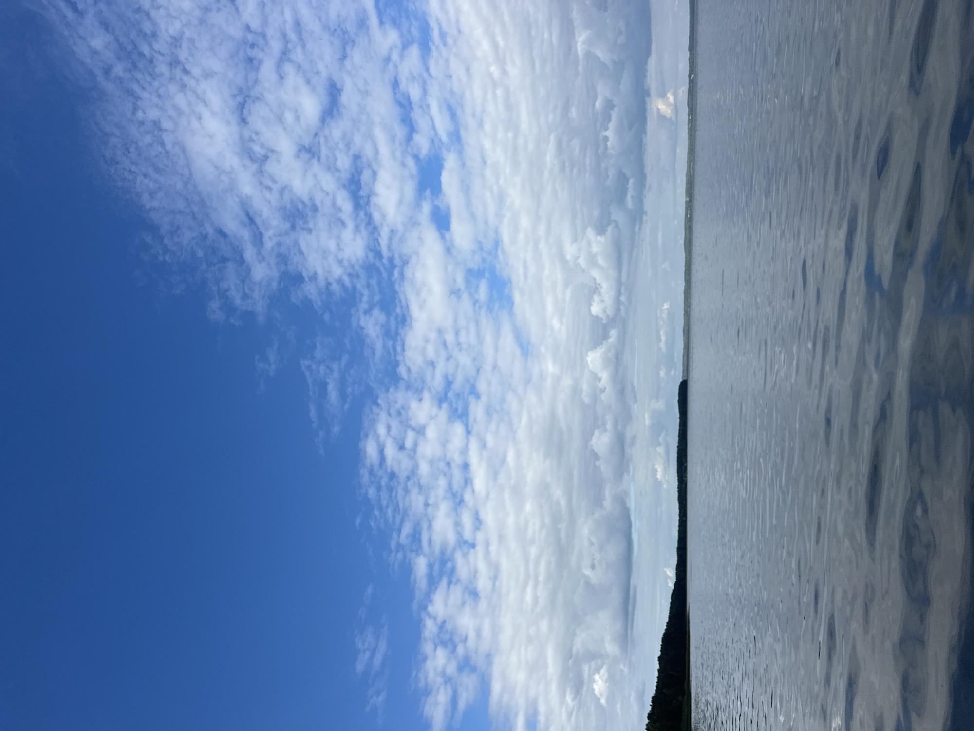 The image shows a calm body of water, likely a lake, with a dark green tree line on the horizon. The sky is a vibrant blue with scattered white clouds, some of which are fluffy and cumulus-like, while others are thinner and wispy. The water's surface has gentle ripples reflecting the sky. The scene evokes a sense of tranquility and natural beauty. The location is in the Нарацкі сельскі Савет, Belarus, a region known for its numerous lakes and picturesque landscapes. The image depicts an outdoor natural environment without any people or man-made structures visible.