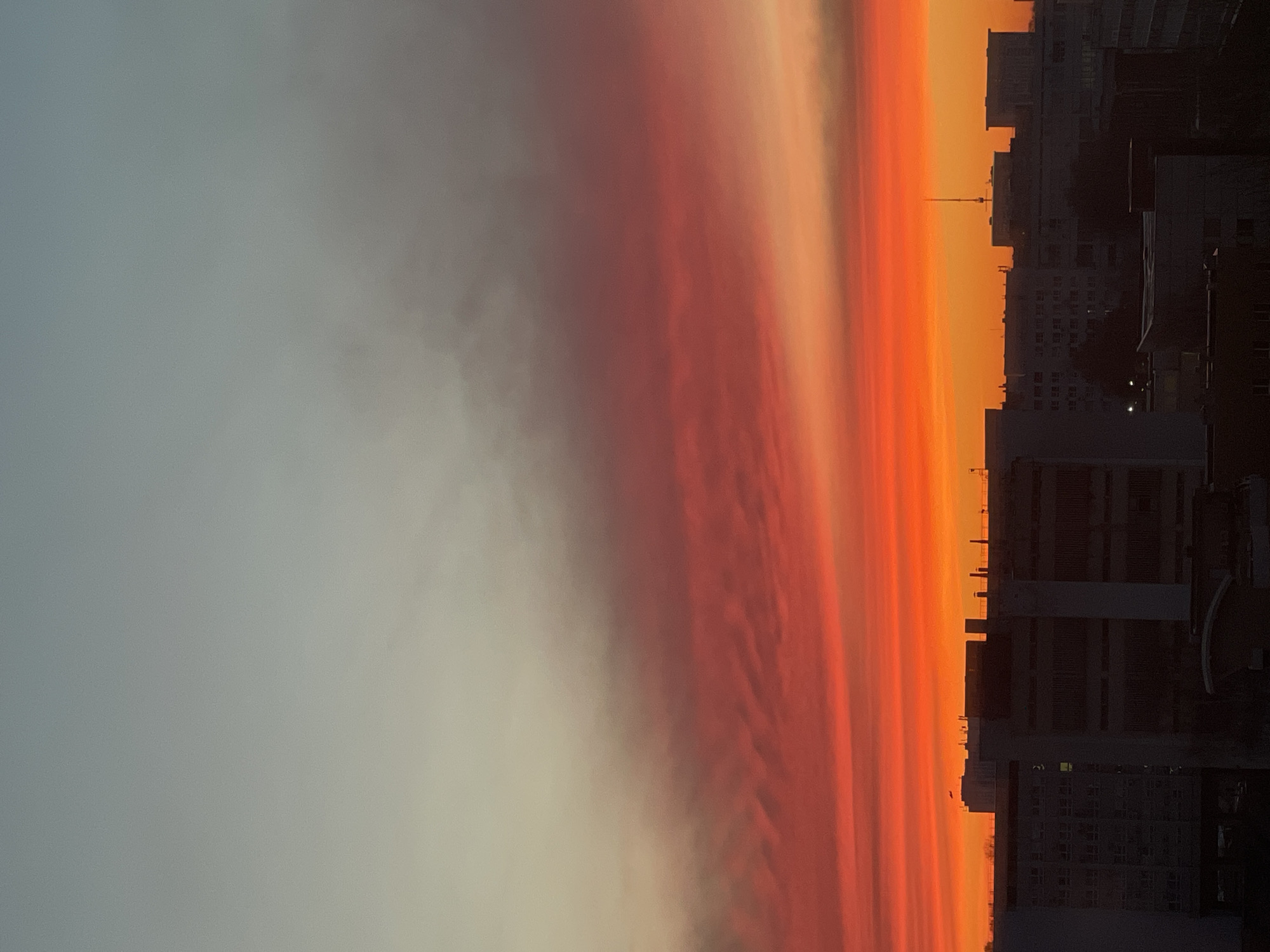 The image depicts a vivid sunset over Izhevsk, Russia. The sky transitions from a pale gray to a gradient of red, orange, and yellow hues. On the right side of the image, silhouettes of buildings and structures are visible against the bright sky. The buildings appear to be of varying heights and architectural styles.